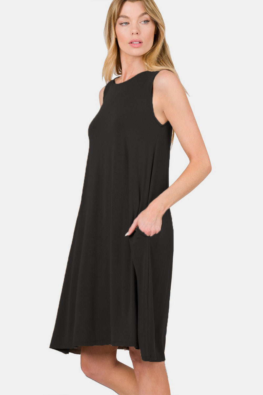 A woman with blonde hair smiles while wearing the Zenana Full Size Sleeveless Flared Dress with Side Pockets in black. She holds one side of the dress with her hand and has her other hand in the pocket, showcasing this versatile wardrobe piece.