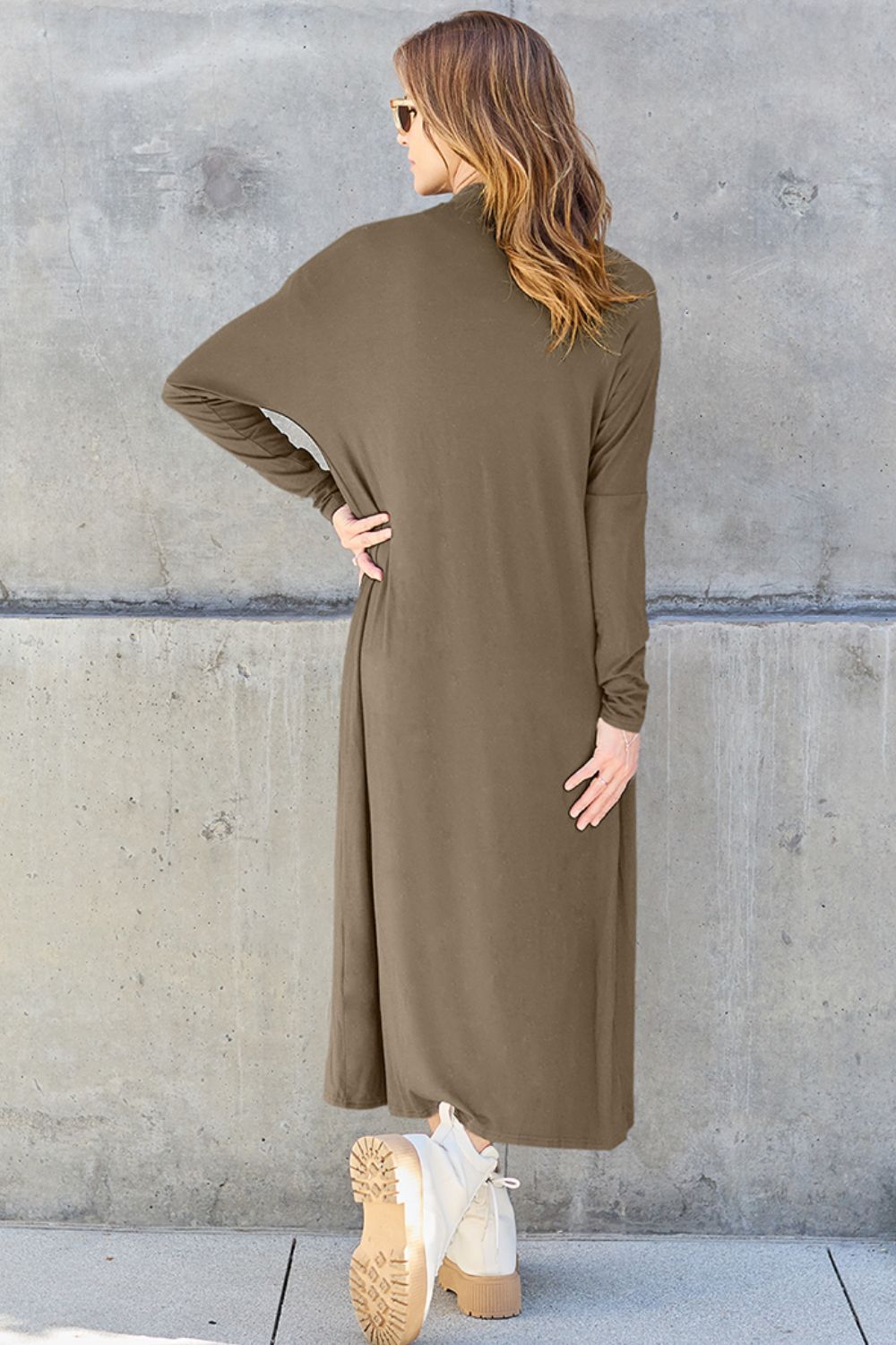 A woman stands against a concrete wall, wearing the Basic Bae Full Size Open Front Long Sleeve Cover Up made from rayon spandex, a white top, blue jeans in her perfect size, and brown shoes with her hands in her pockets. Machine wash cold for best results.