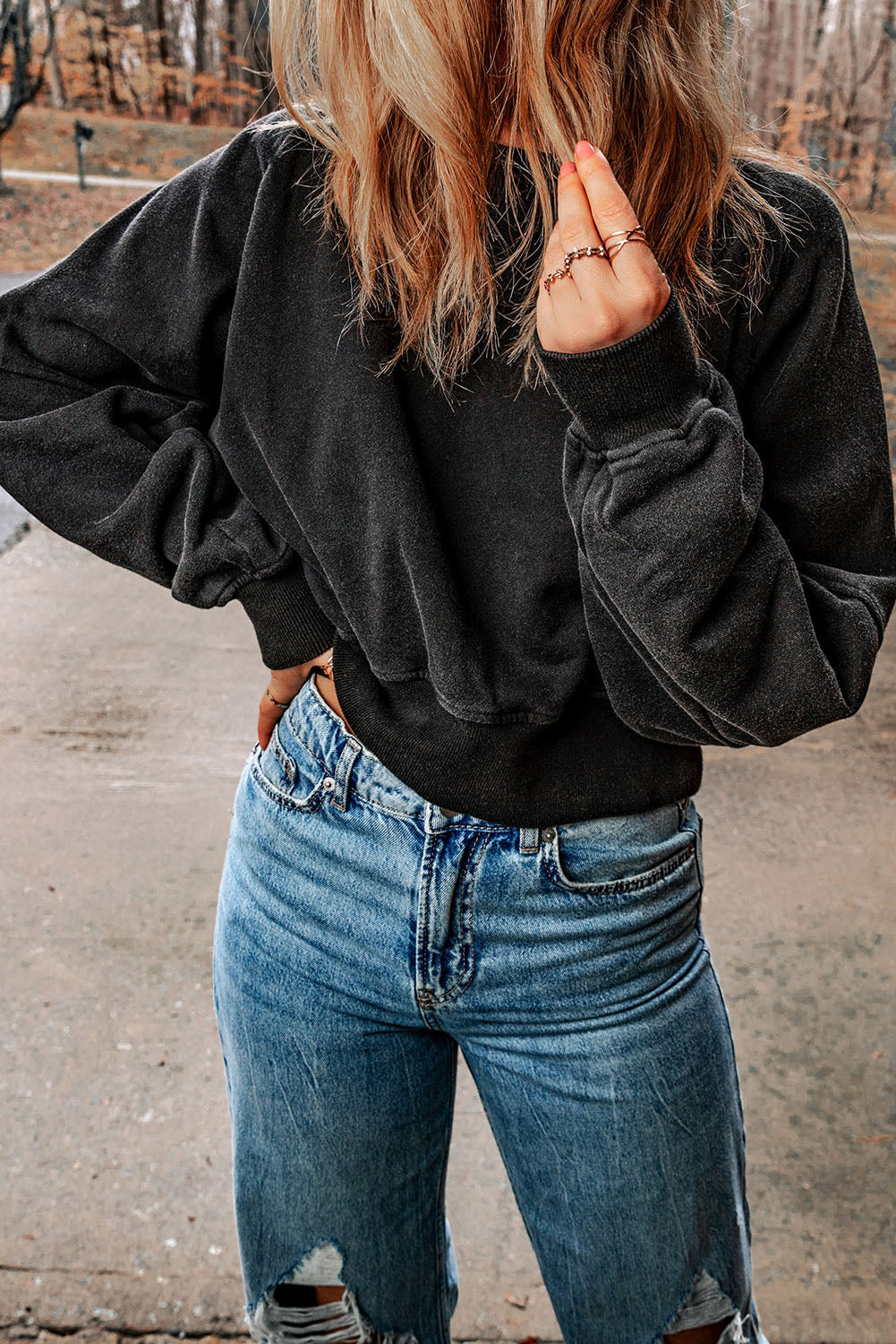 A person with wavy, shoulder-length hair wearing a Black Acid Wash V-shape Open Back Sweatshirt and distressed blue jeans stands outside on a paved surface.