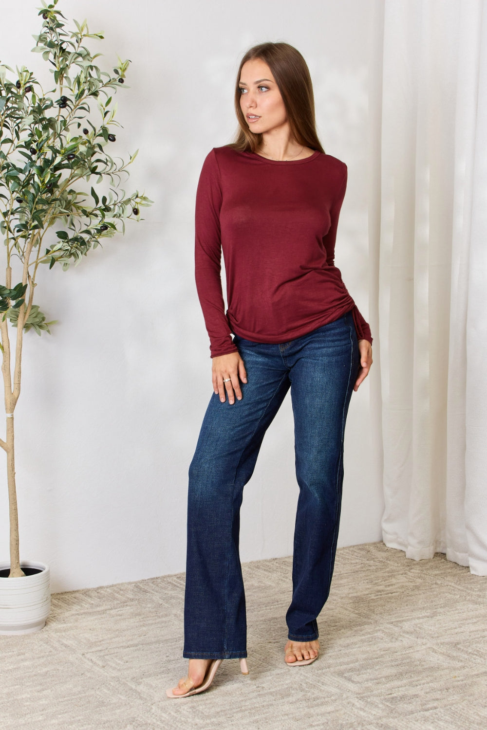 A woman stands in front of a plain background and a small tree, wearing the Culture Code Full Size Drawstring Round Neck Long Sleeve Top in maroon and blue jeans.