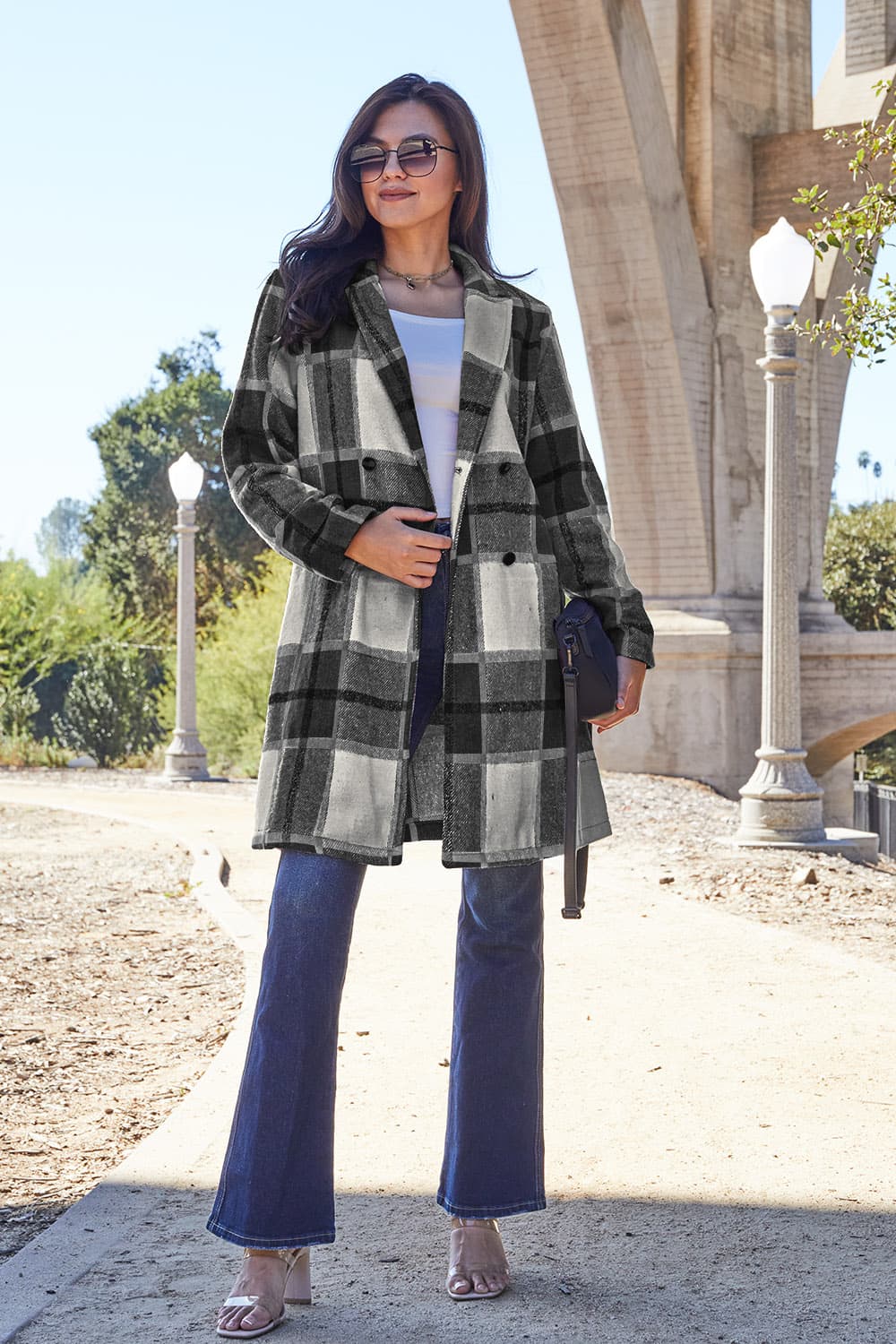 A woman stands on a sidewalk wearing the Double Take Full Size Plaid Button Up Lapel Collar Coat in pink and white over a white top and blue jeans, with her right hand lightly touching her hair. The polyester fabric of the coat makes it durable and perfect for an easy machine wash cold.