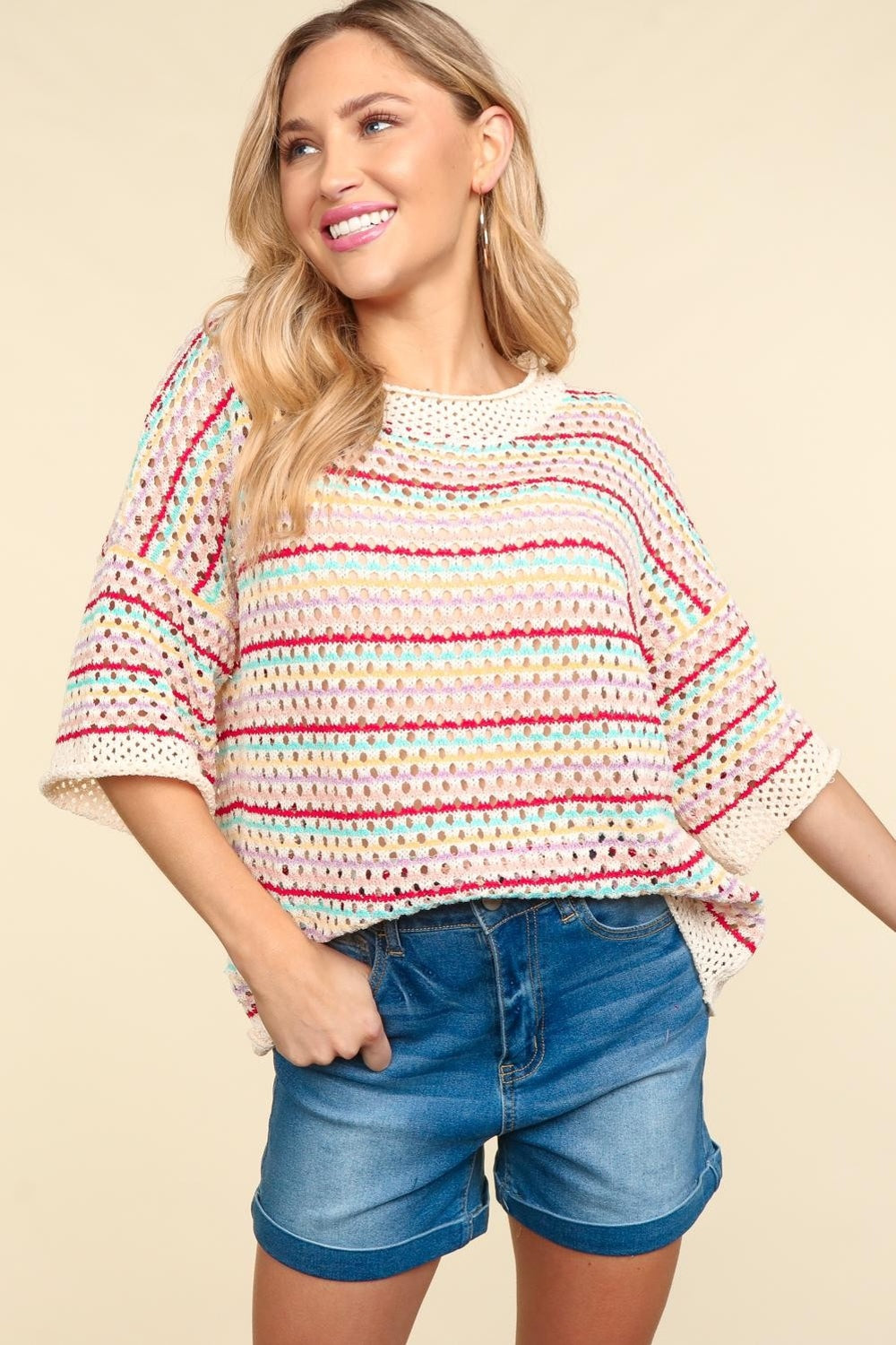 A woman stands smiling against a beige background, wearing the Haptics Striped Crochet Drop Shoulder Knit Top paired with denim shorts.