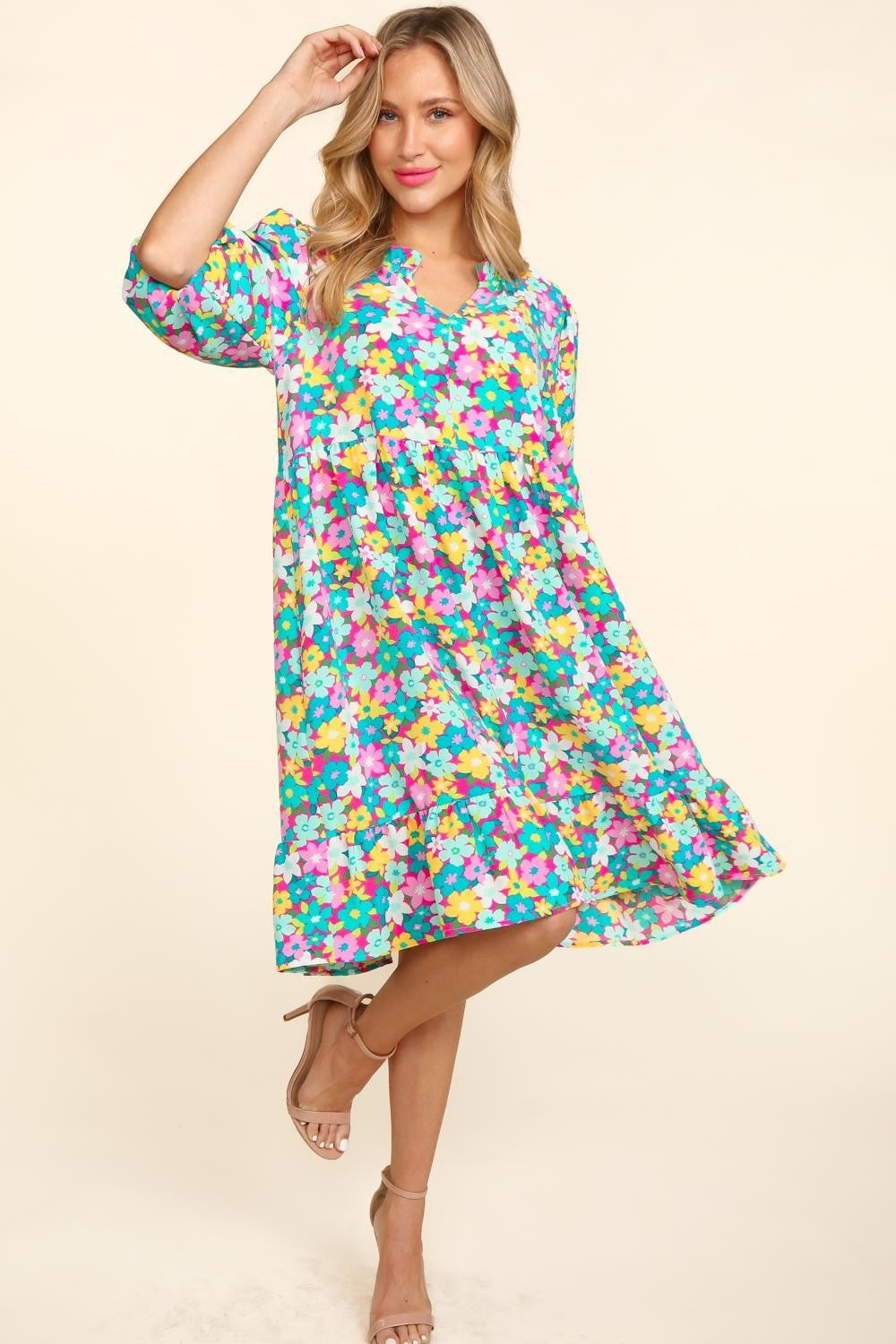 A woman in a Haptics Bubble Sleeve Floral Ruffled Dress stands against a plain background, with one hand holding her dress and the other by her side. She has long, wavy hair and is wearing open-toed shoes, perfect for any garden party.