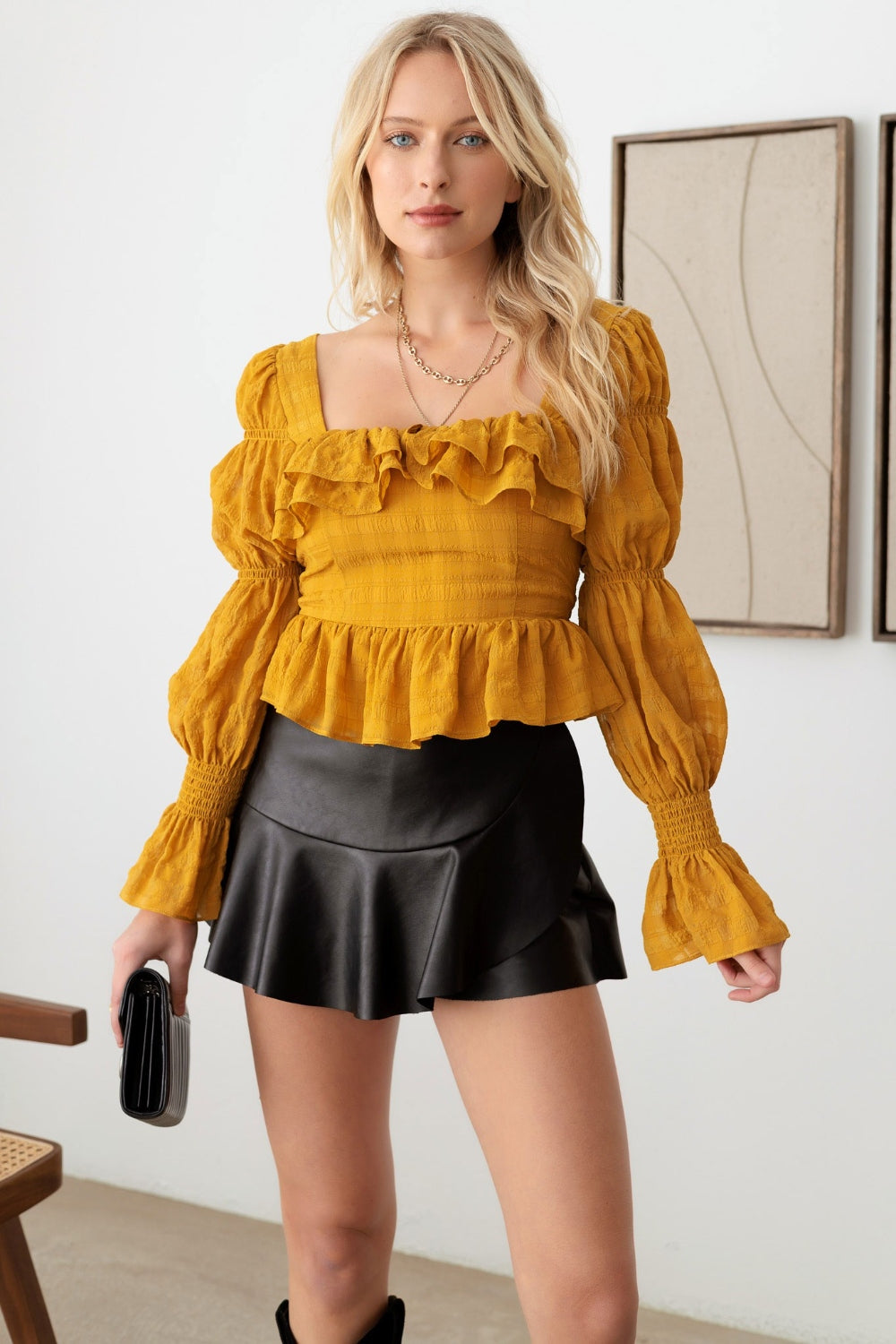 A woman with long blonde hair is wearing the Mustard Seed Boho Shirred Smocked Peplum Long Sleeve Top, a black skirt, and holding a black purse. She is standing in front of a white wall adorned with framed art.