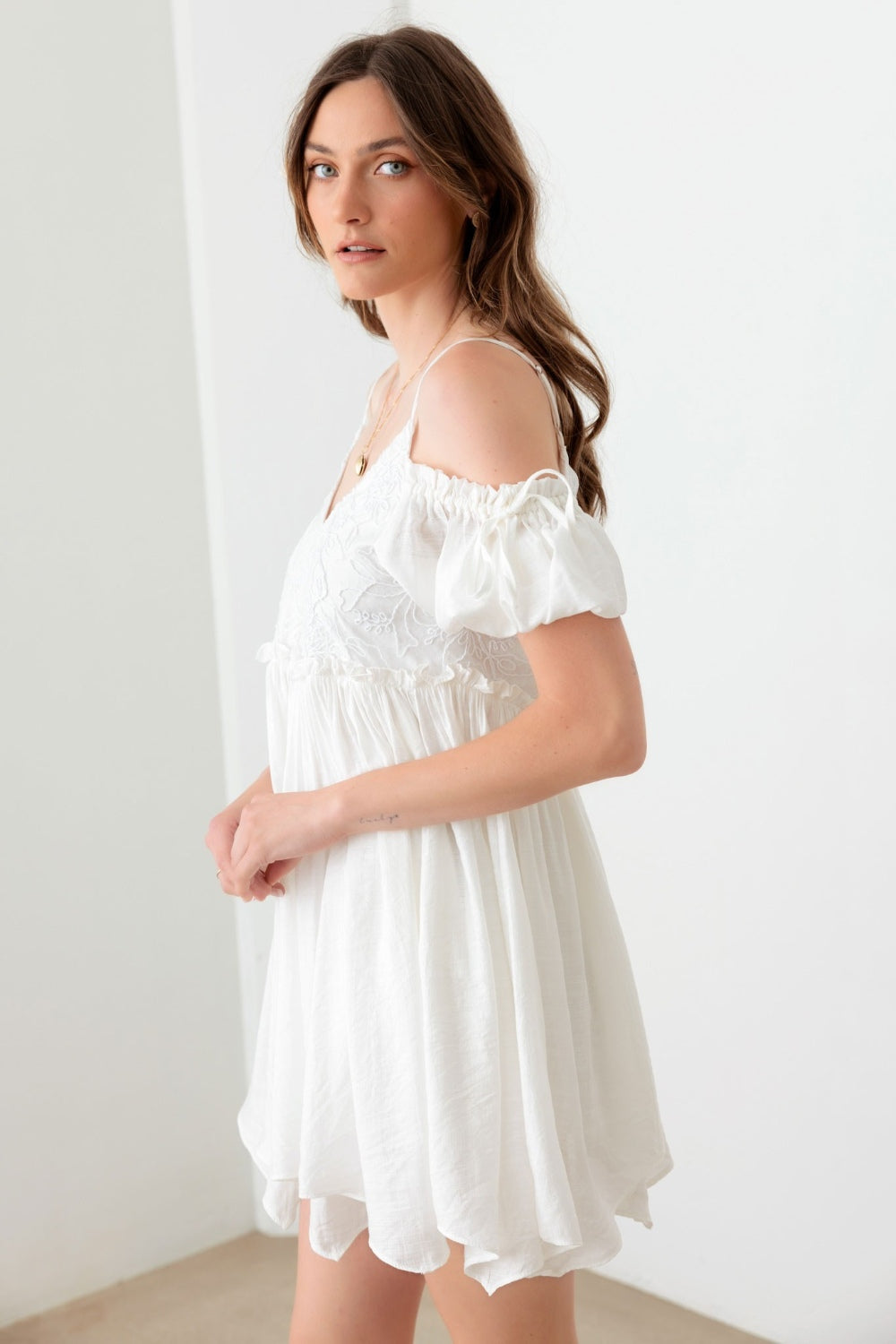 A woman poses in a boho-inspired, embroidered floral shirred cold shoulder mini dress by Mustard Seed. She accessorizes with a gold necklace and maintains a neutral expression against the backdrop of a plain white wall.