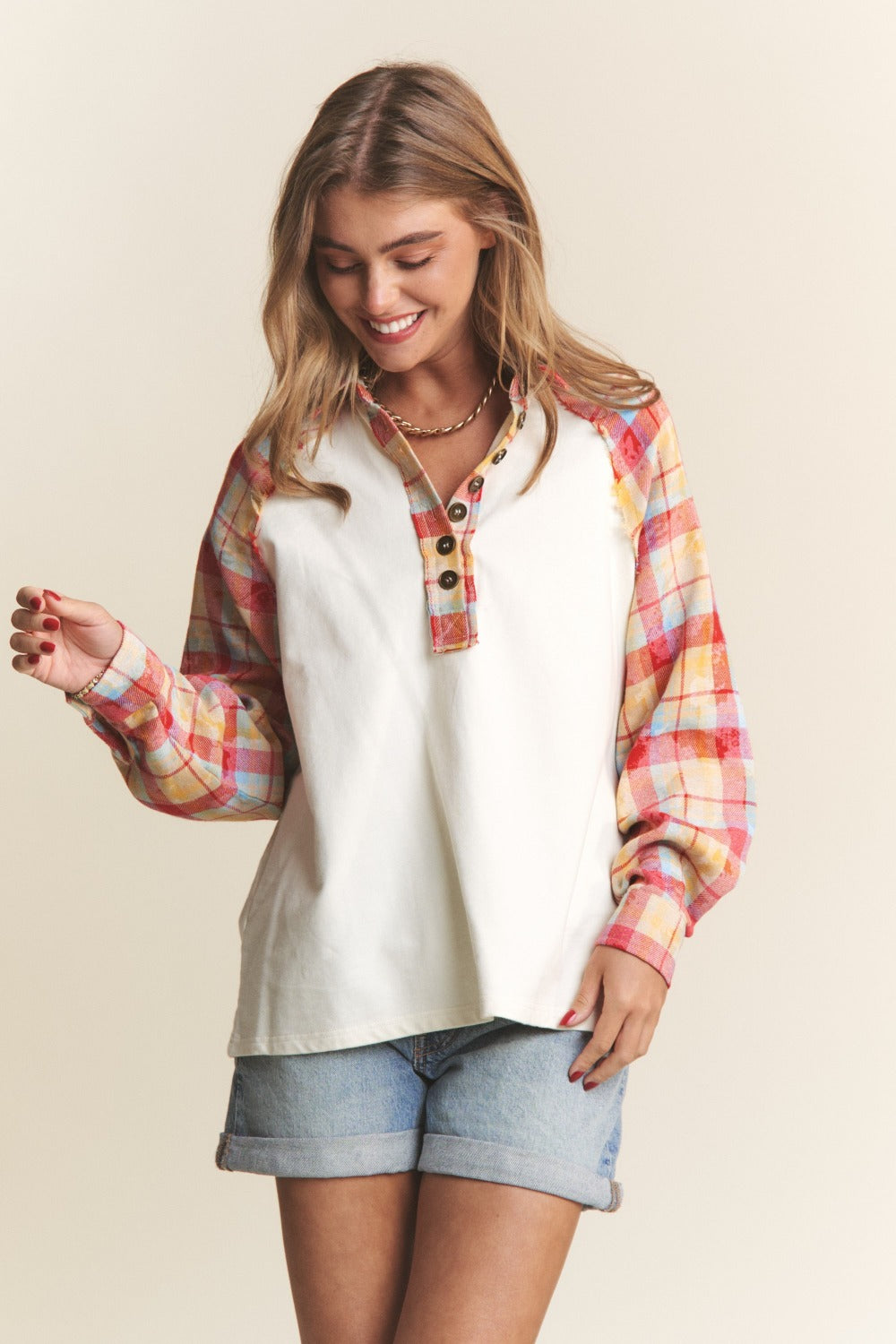 A woman wearing the J.NNA Plaid Contrast Button Down Henley Top—a stylish white pullover shirt with red and yellow plaid sleeves—and denim shorts, stands against a plain background.