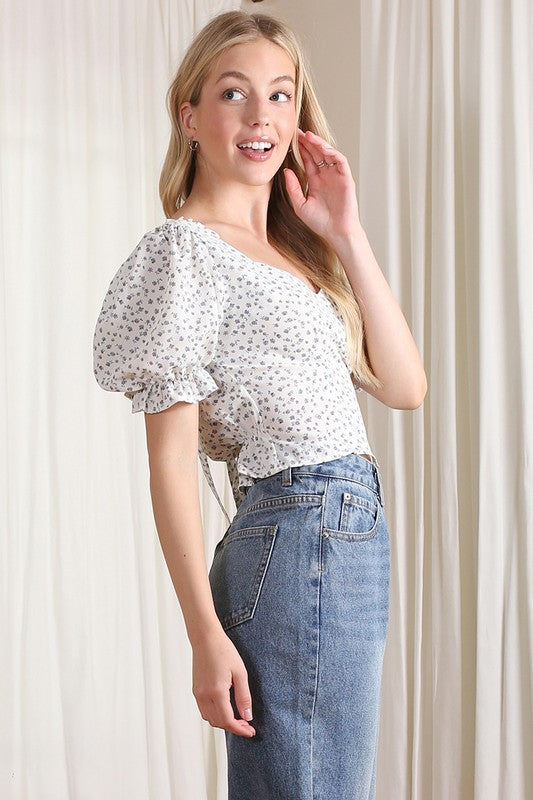 A person wearing the SS puff sleeve top, featuring a blue floral print, pairs it with light jeans while standing on grass beside potted plants, against a white wall backdrop.