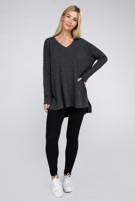 A woman with long, blonde hair is wearing a loose-fitting, brown Dolman Long Sleeve V-Neck Side Slit Hi-Low Hem Top and black pants. She poses with one hand on her hip and a neutral facial expression against a light grey background.