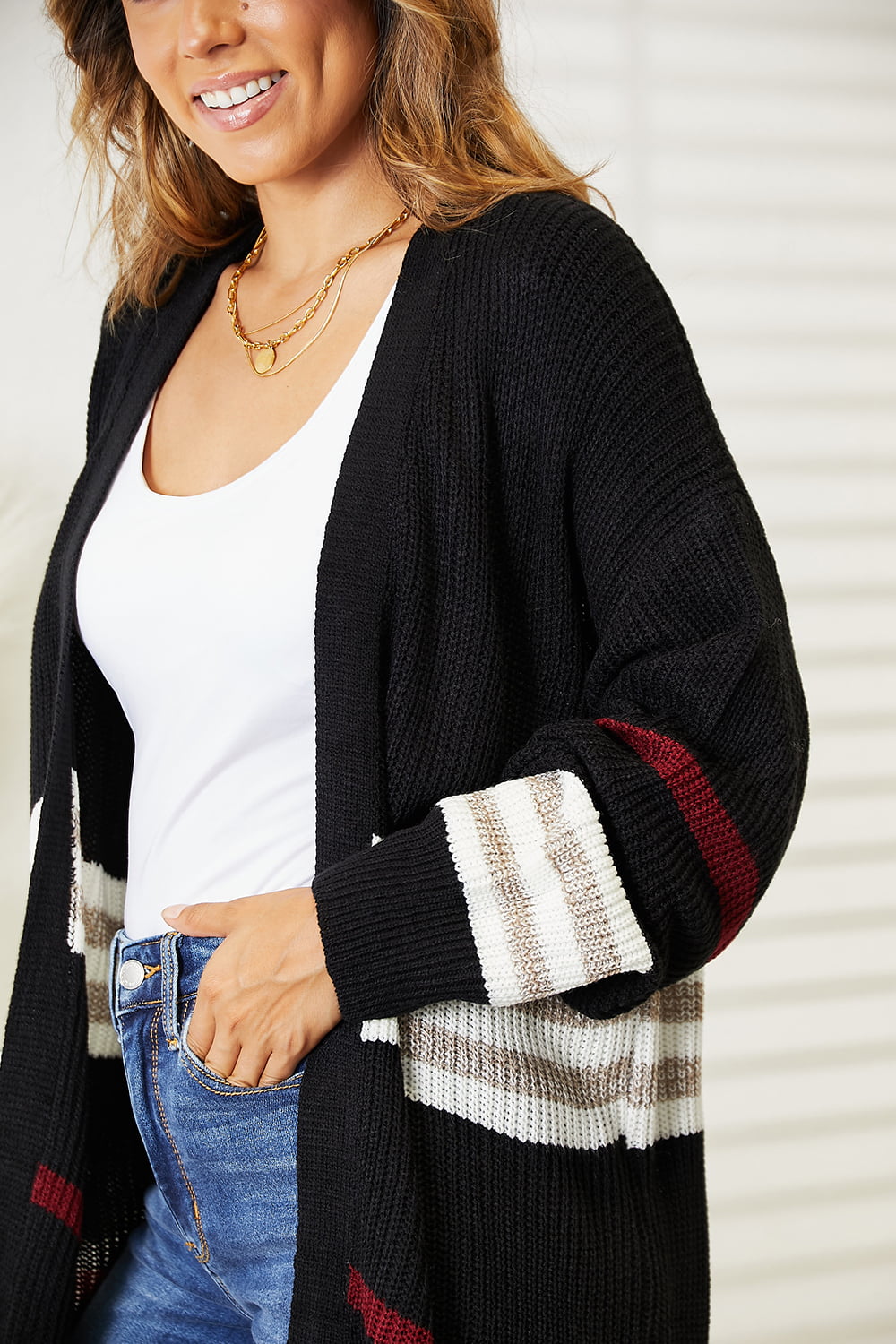 A person wearing a Perfee Striped Rib-Knit Drop Shoulder Open Front Cardigan with white and red accents, a white top, blue jeans, and layered necklaces stands next to a decorative table.