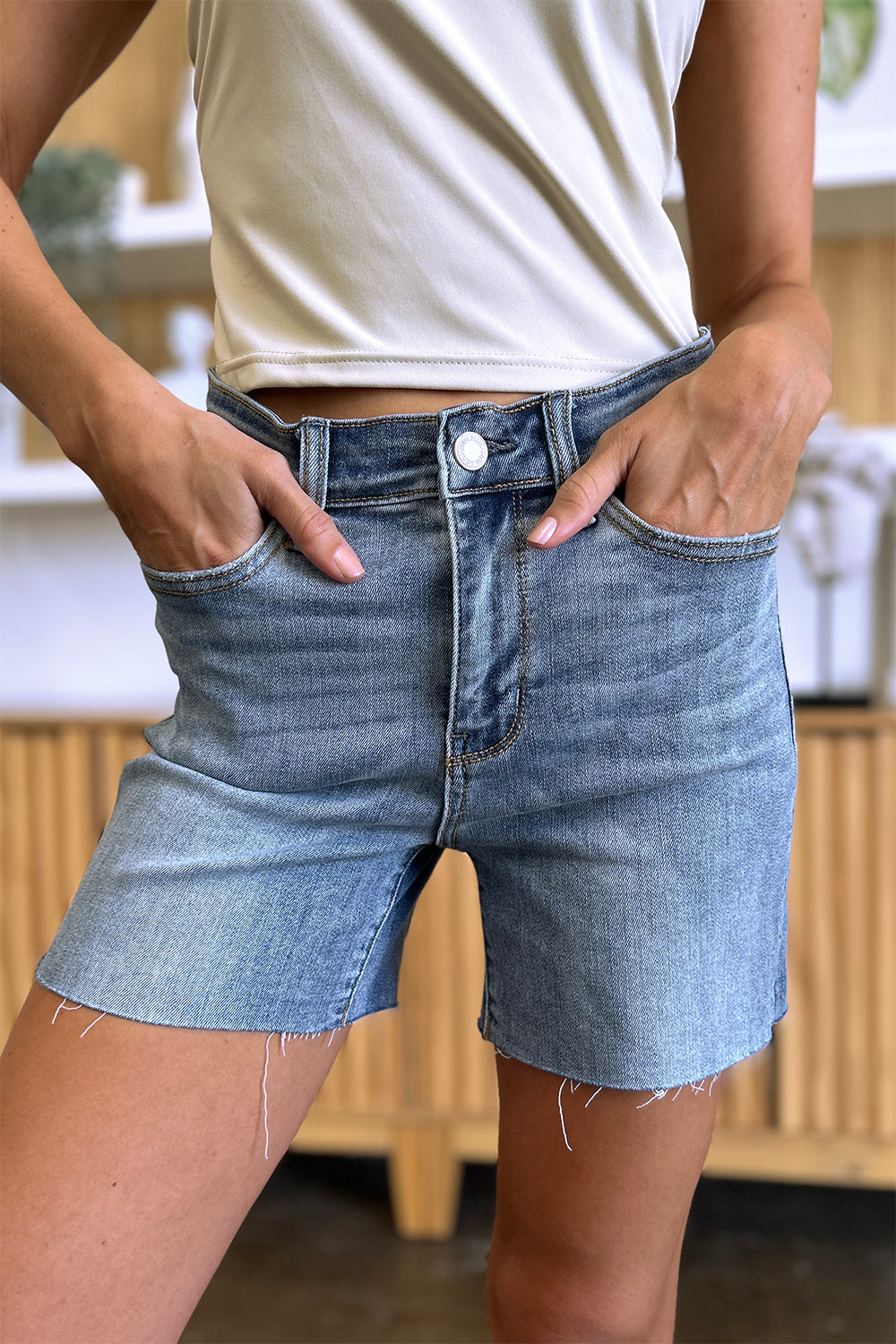 Side view of a person wearing a white shirt and Judy Blue Full Size High Waist Raw Hem Denim Shorts, standing indoors near a wooden cabinet. Summer fashion vibes radiate through the casual ensemble.