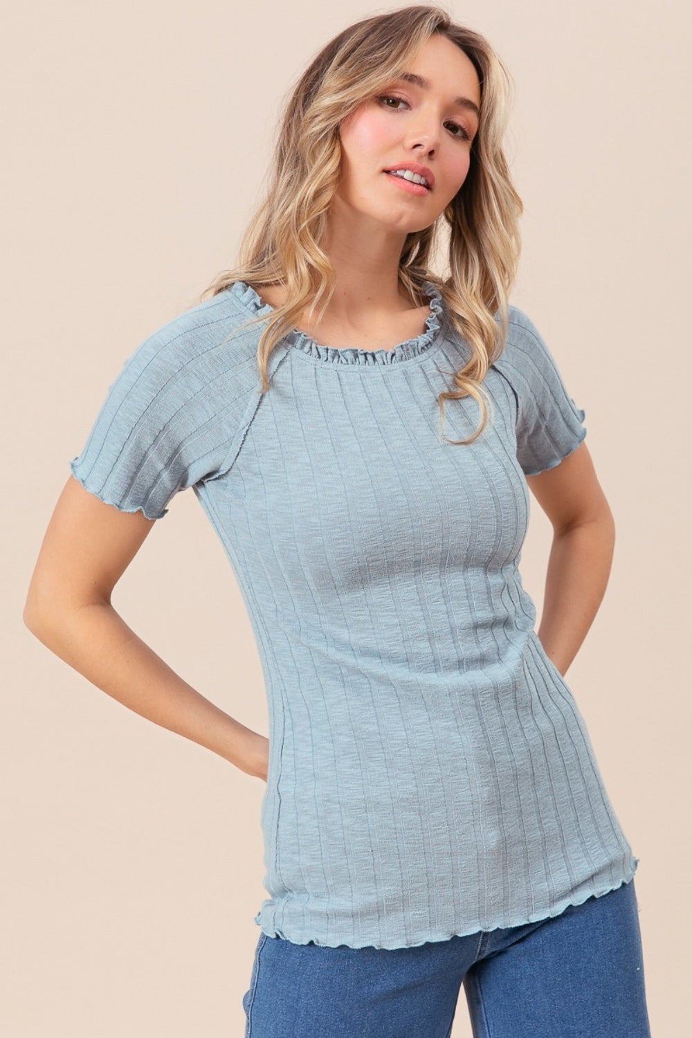 A person wearing the BiBi Ruffle Round Neck Merrow Edge Rib Top, featuring short sleeves and a light blue hue, stands against a neutral background. They have long, wavy hair, with tattoos adorning their arm.