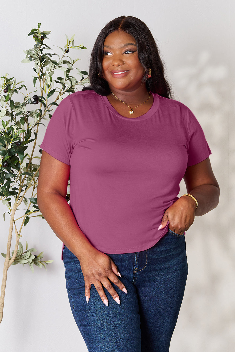 A woman with long hair, smiling with her right hand in her pocket and standing next to a plant, is wearing the Basic Bae Full Size Round Neck Short Sleeve T-Shirt in black along with beige pants made of rayon spandex.