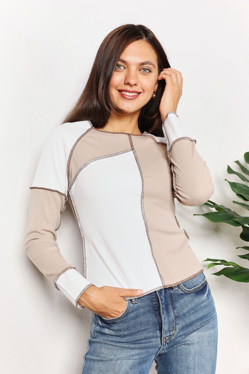 A woman with long brown hair is wearing the "Double Take Color Block Exposed Seam Top" in beige and white, paired with jeans. She stands with one hand in her pocket and the other touching her hair, smiling. A plant is visible in the background.