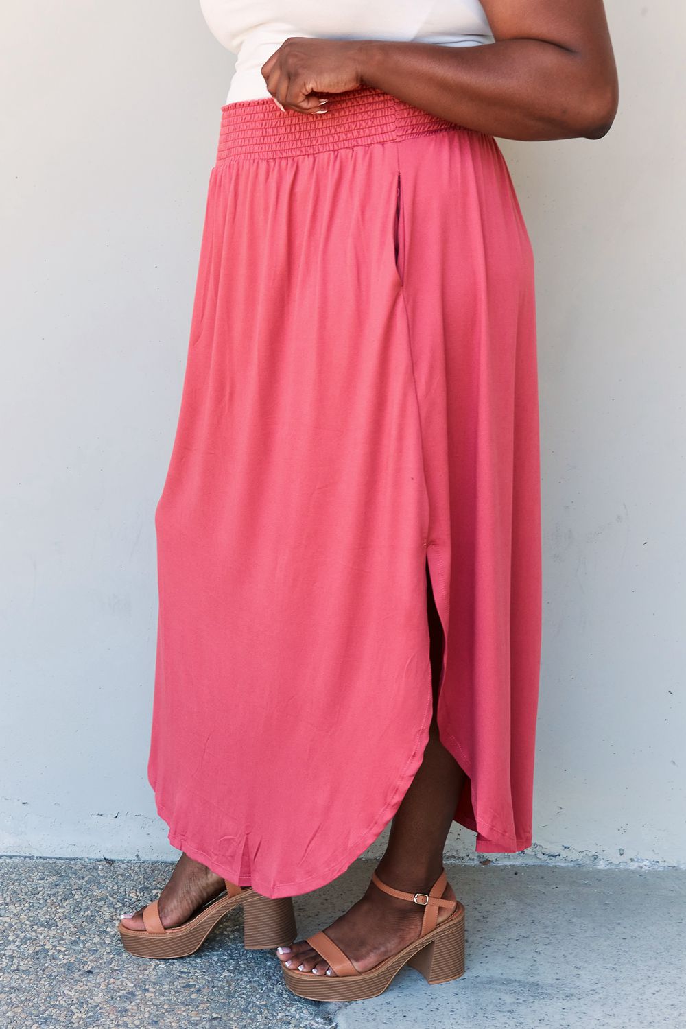 A person wearing a white top and the Doublju Comfort Princess Full Size High Waist Scoop Hem Maxi Skirt in an elegant coral color, paired with tan sandals, stands in front of a plain wall.