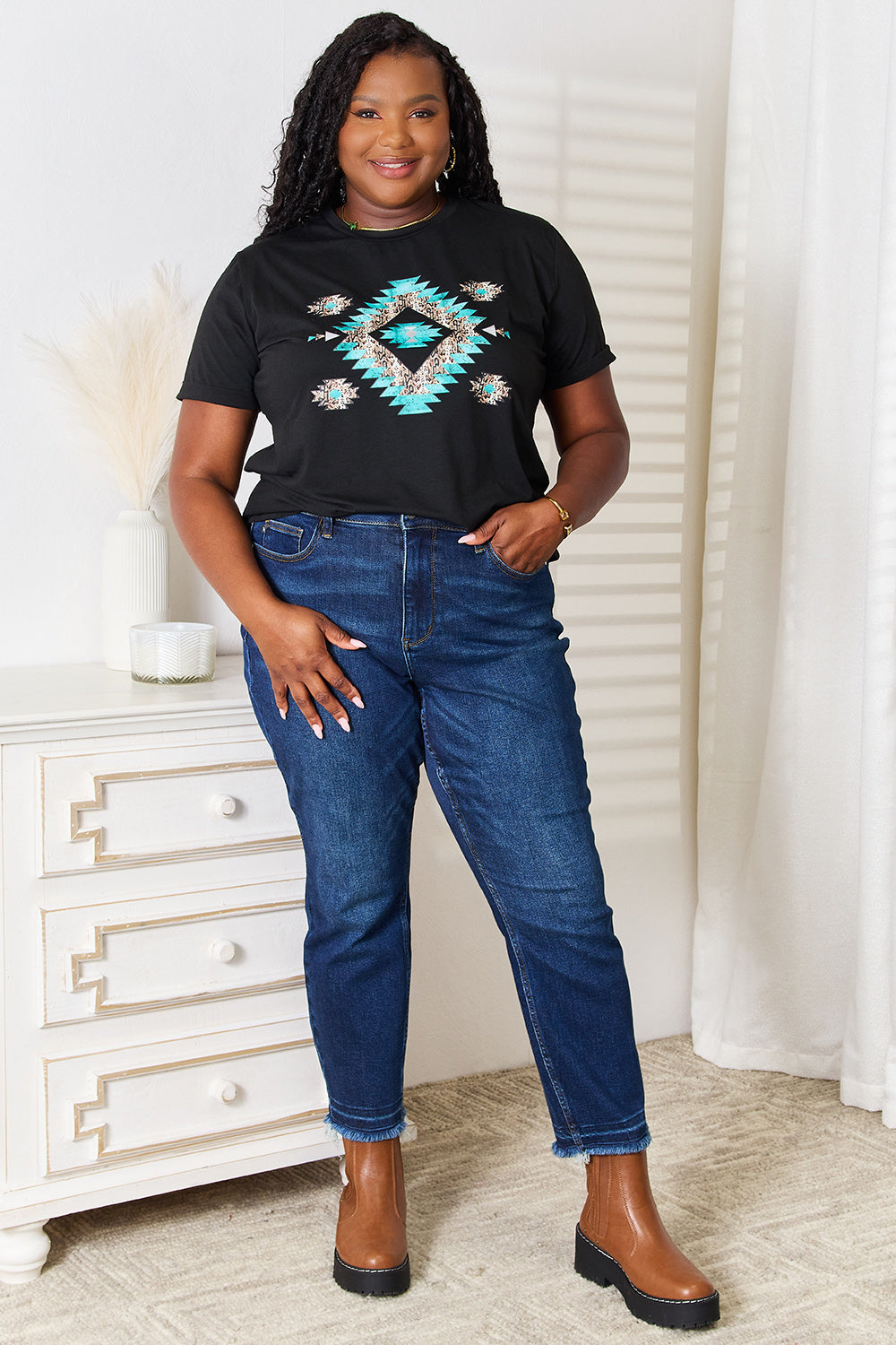 A woman stands indoors wearing the Simply Love Graphic Short Sleeve T-Shirt, a stylish black tee featuring a striking blue and white geometric pattern. Made from comfortable fabric, she has one hand on her hip and is smiling.