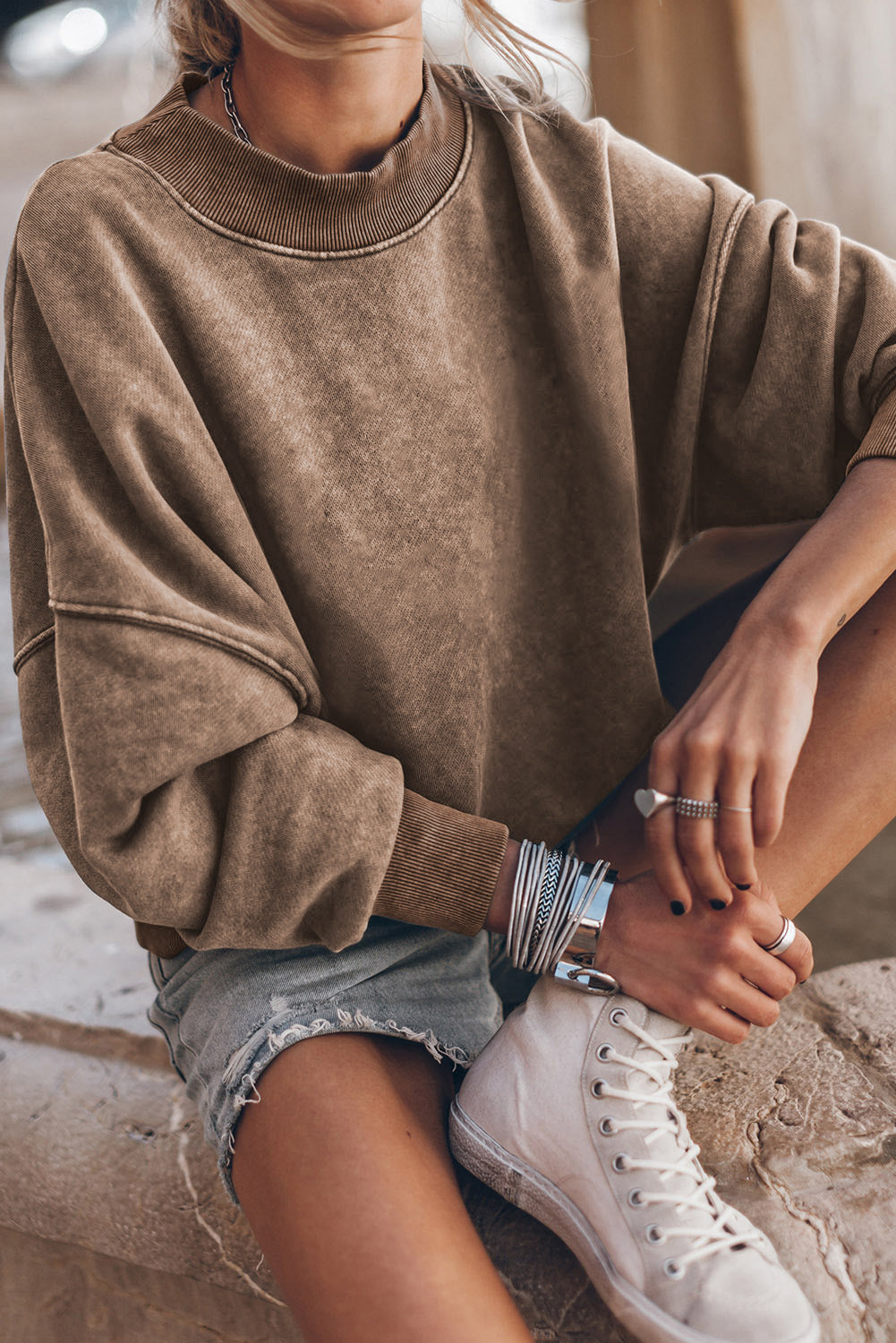 A person is seen wearing a brown Drop Shoulder Crew Neck Pullover Sweatshirt with rolled-up sleeves, paired with light blue jeans and standing with one hand in their pocket. Multiple bracelets are visible on their wrist while the background appears blurred.