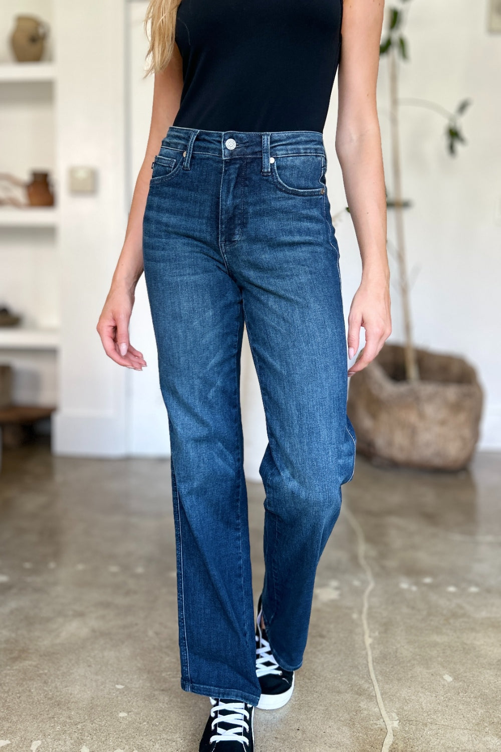 A person wearing a black top and Judy Blue Full Size Tummy Control Straight Jeans stands indoors with one hand on their hip. Their shoes are black with white soles. A plant and shelves are in the background.