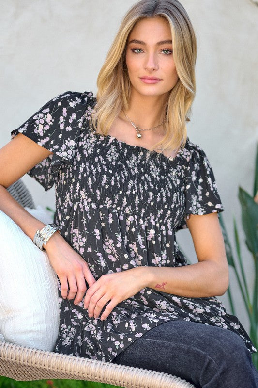 A woman stands outdoors donning a Floral Printed V-Neck Ruffle Top paired with jeans, set against a neutral background, with plants in the foreground, highlighting her fashion-forward wardrobe.