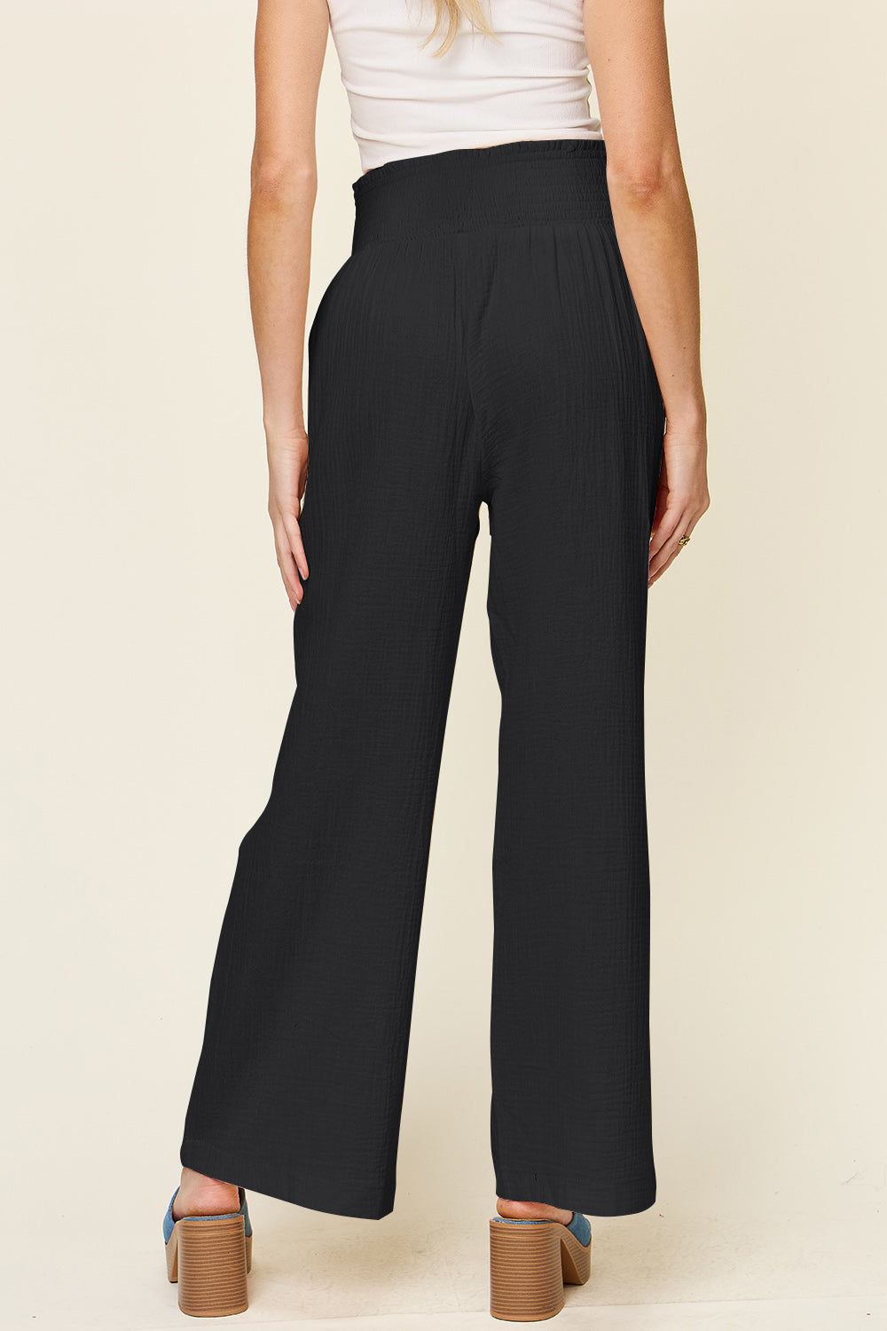 A person wearing Double Take Full Size Texture Smocked Waist Wide Leg Pants in black and a white 100% cotton top is standing with one hand on their hip. They are also wearing blue open-toe sandals.