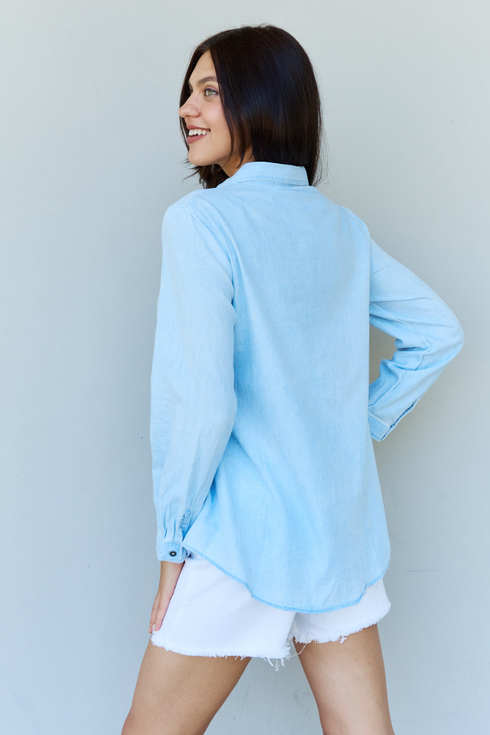 A woman wearing the Doublju Blue Jean Baby Denim Button Down Shirt Top in Light Blue, paired with white ripped shorts, strikes a pose with one hand in her pocket against a plain background, highlighting the classic button-up silhouette.