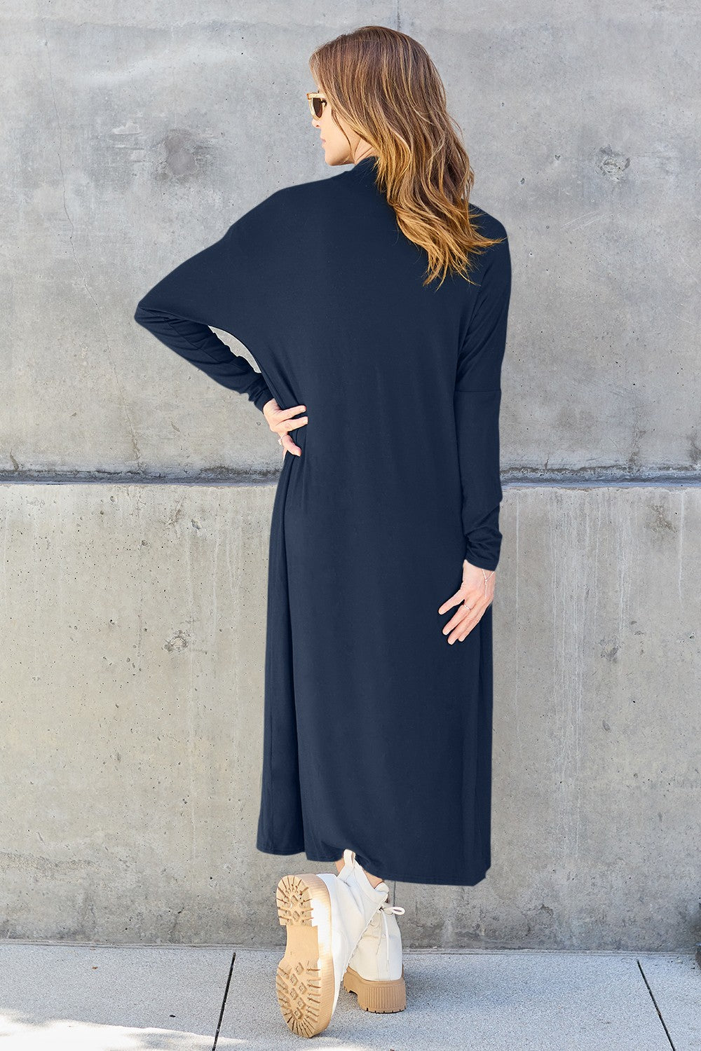 A woman stands against a concrete wall, wearing the Basic Bae Full Size Open Front Long Sleeve Cover Up made from rayon spandex, a white top, blue jeans in her perfect size, and brown shoes with her hands in her pockets. Machine wash cold for best results.