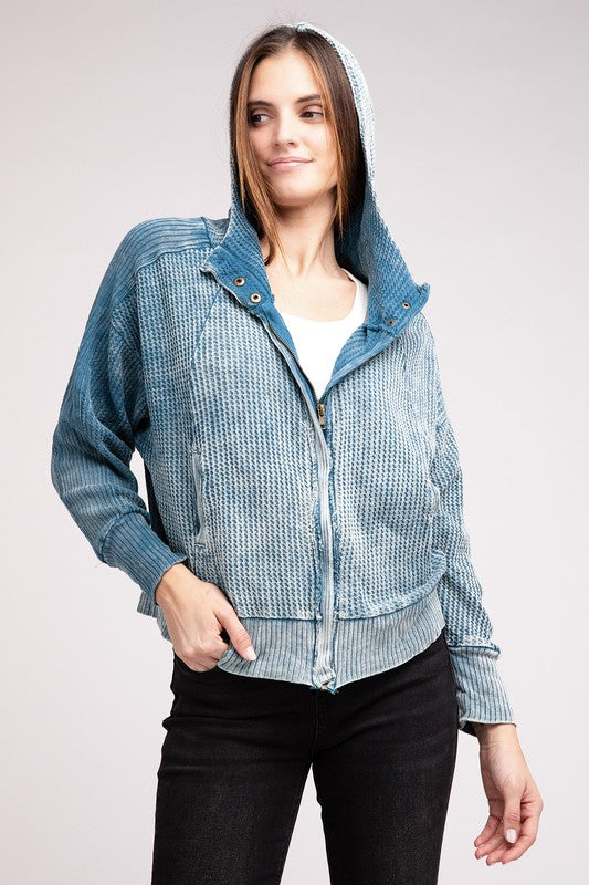 Woman in a Acid Wash Cotton Waffle Hooded Zip-Up Jacket with pockets and black pants, smiling and posing with hands on hips, against a light gray background.