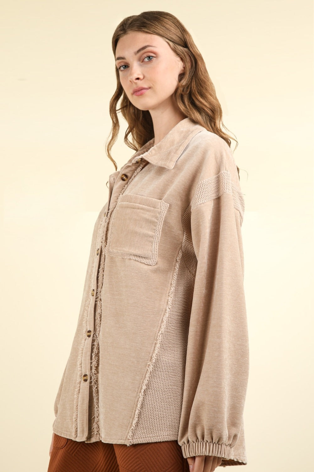 A woman with long wavy hair wears the VERY J Mixed Media Button Down Raw Hem Shacket featuring a drop shoulder design over a brown outfit. She faces the camera with one hand holding the lapel of her shacket against a plain beige background.