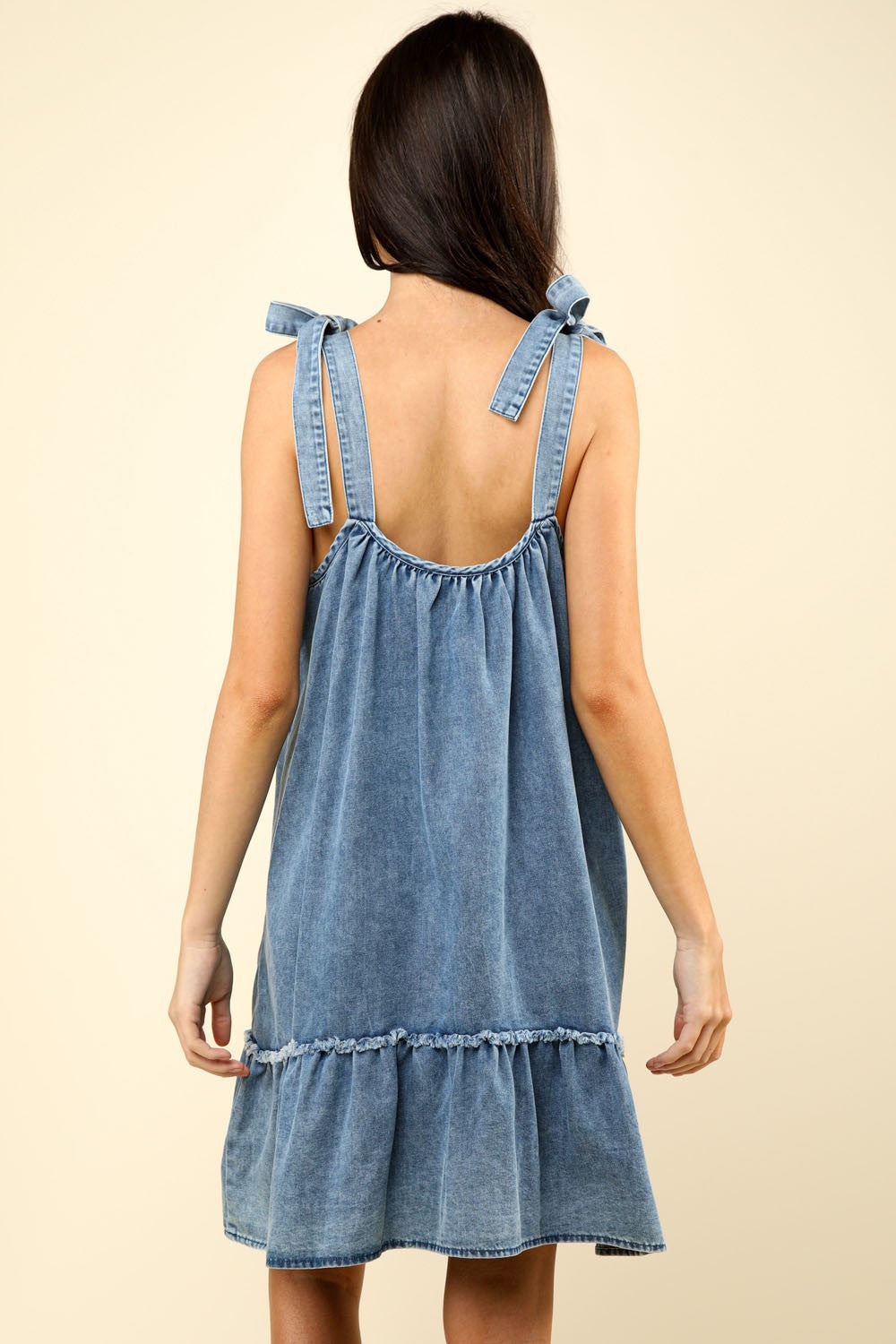 A woman wearing the VERY J Shoulder Tie Washed Denim Mini Dress, featuring a stylish ruffled hem and tied shoulder straps, casually holds sunglasses on her head against a beige background.