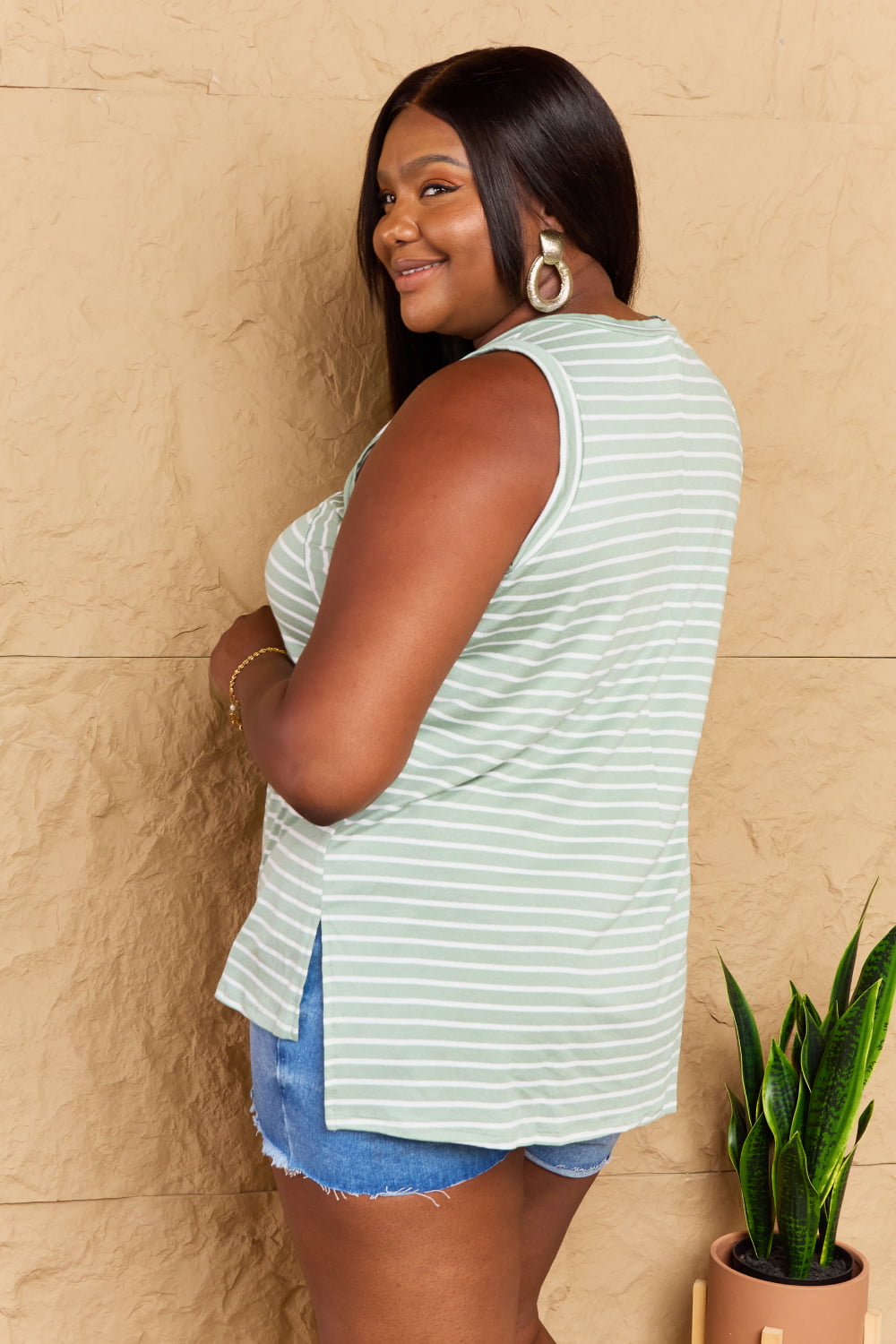 A person wearing the Doublju Full Size Striped Sleeveless V-Neck Top paired with denim shorts stands against a textured beige wall, smiling and posing with one hand on their hip.