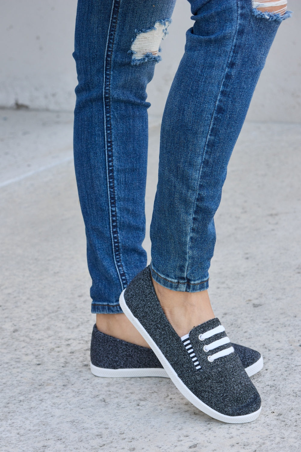 A person wearing ripped blue jeans and the fashionable Forever Link Round Toe Slip-On Flat Sneakers with white elastic bands.