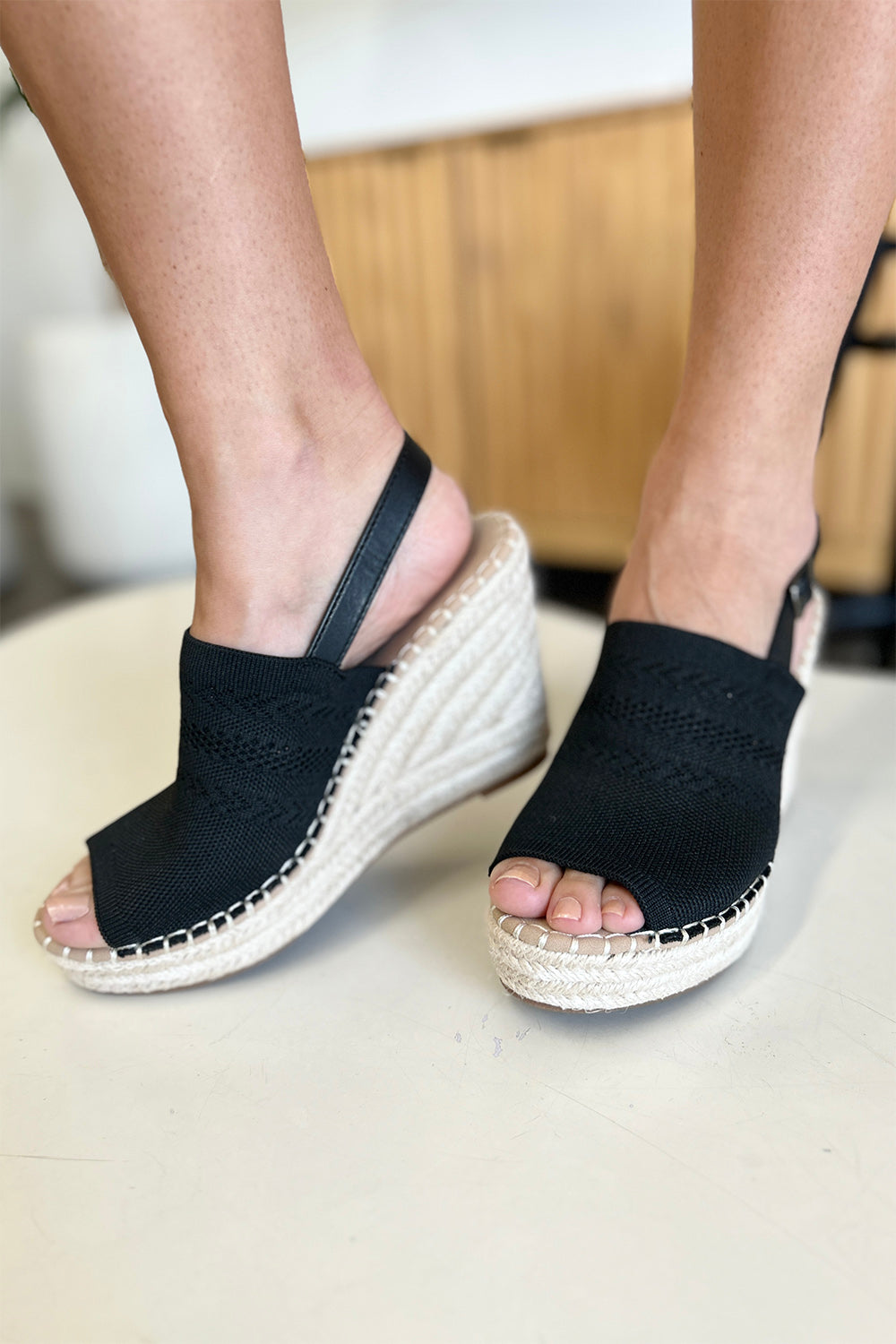 Close-up of a person's feet wearing chic black Forever Link Peep Toe High Heel Sandals with a braided espadrille base and adjustable heel straps. The background is blurred, making these sandals the stylish choice for any outfit.