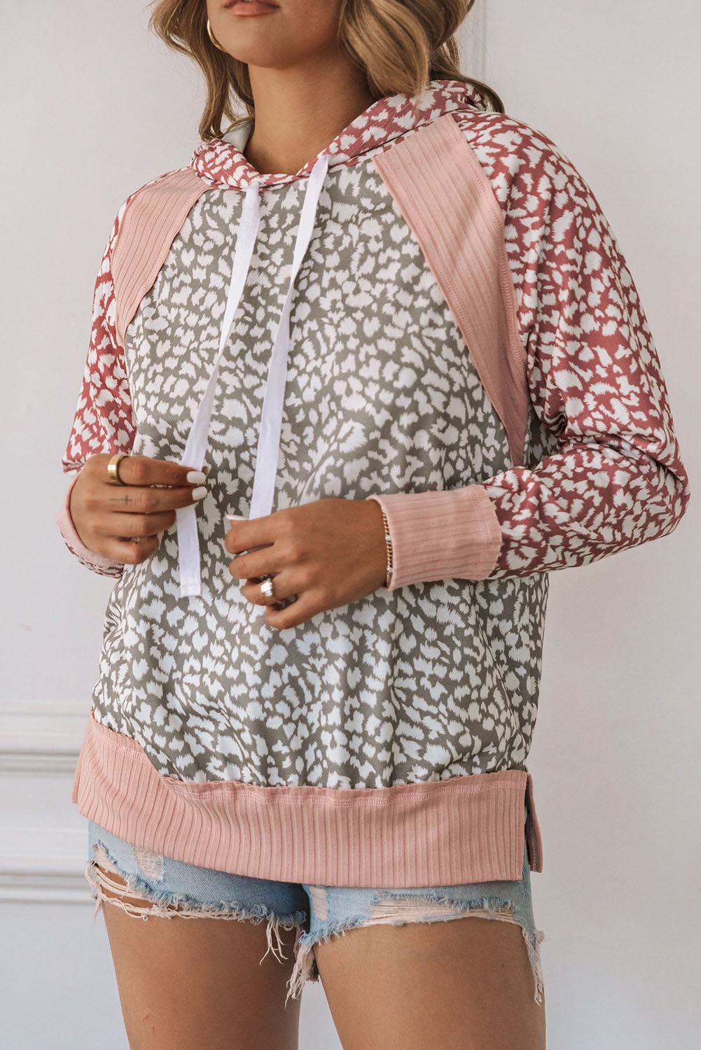 A person with long, wavy hair is facing away, wearing a Pink Leopard Long Sleeve Hooded Sweatshirt crafted from soft and warm fabric with a mix of pink and gray patterns, and light blue denim shorts.