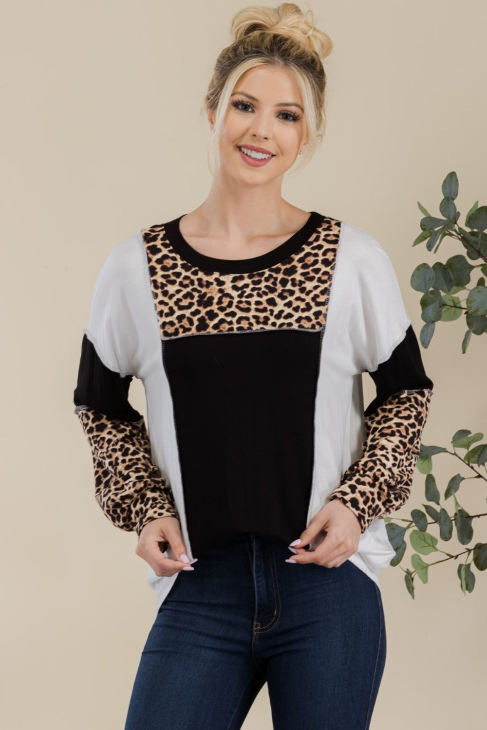 A woman stands in front of a simple background with a plant, wearing the Celeste Full Size Leopard Color Block Exposed Seam T-Shirt, which boasts leopard print cuffs and striking black and white color blocks.