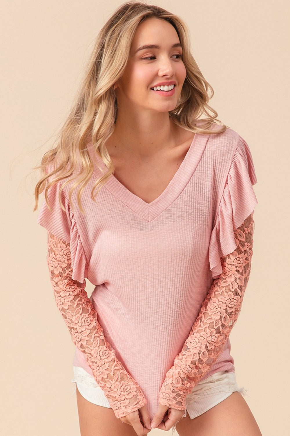 A woman with long blonde hair, smiling and looking to the side, is wearing a pink BiBi Ruffled Lace Sleeve Rib Knit Top paired with white shorts.