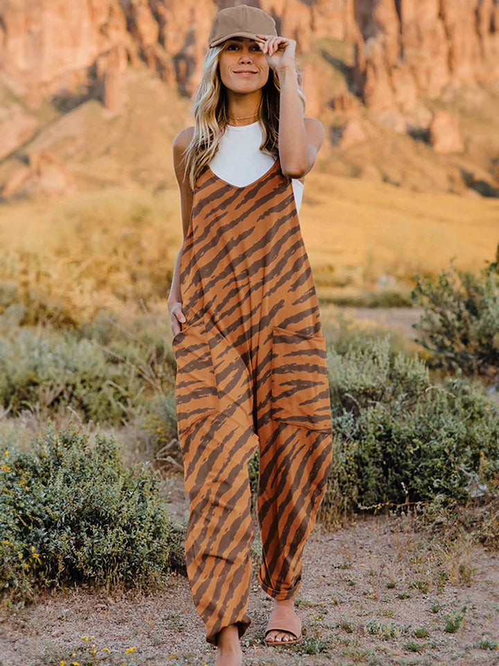 A person wearing a casual white V-neck tank top and a Full Size Printed V-Neck Sleeveless Jumpsuit with a brown animal print zebra-stripe pattern stands outdoors with one hand on their hat and the other in their pocket, set against a rocky landscape backdrop.