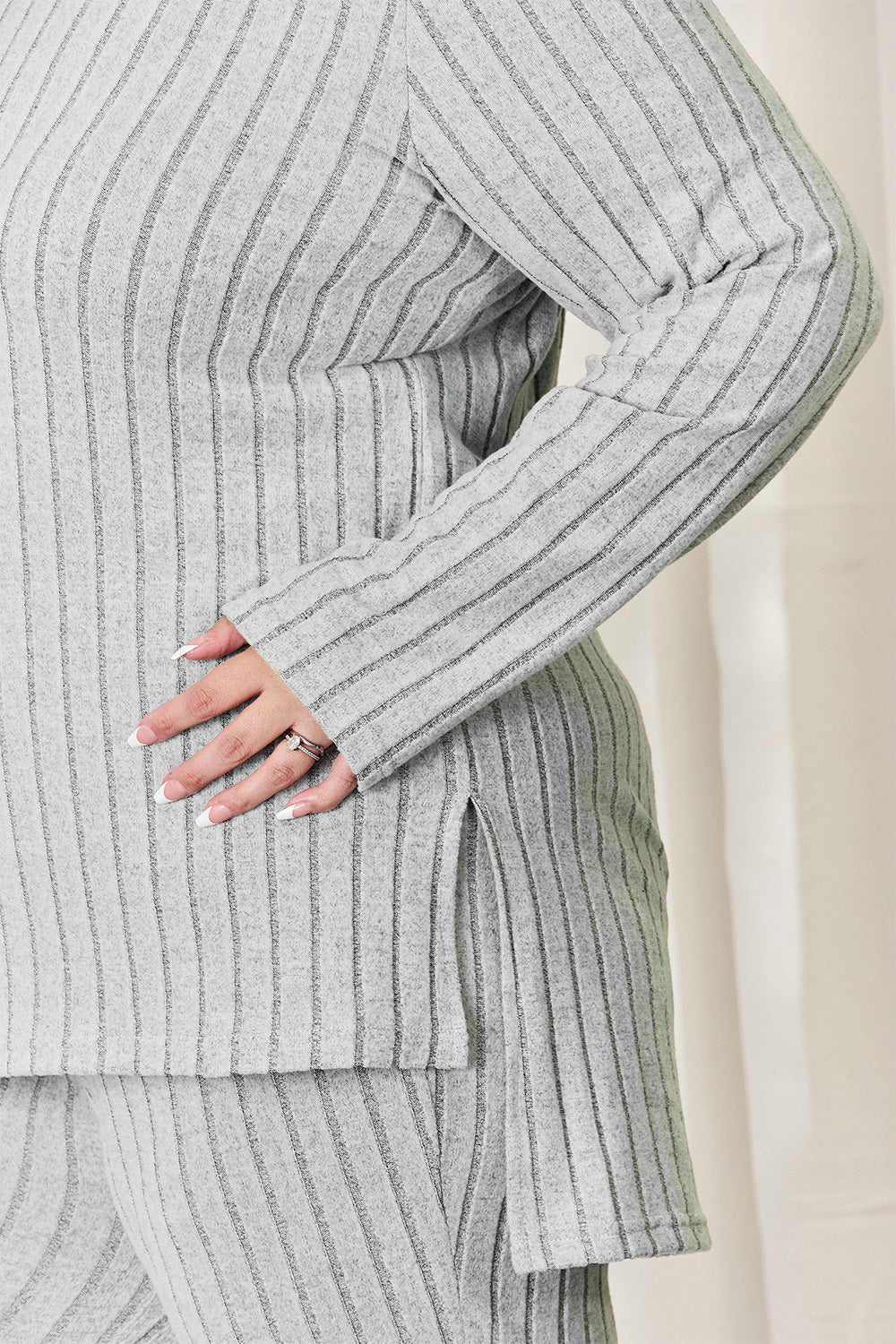 A person wearing the Basic Bae Full Size Ribbed High-Low Top and Wide Leg Pants Set in light gray, made from stretchy material, stands indoors in front of a white curtain.