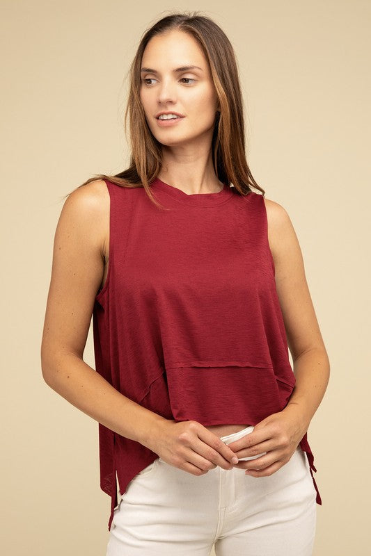 Wearing the Shark Bite Side Slit Short Sleeveless Top in maroon paired with white pants, a model is set against a beige background, exemplifying an effortlessly chic and casual wardrobe.