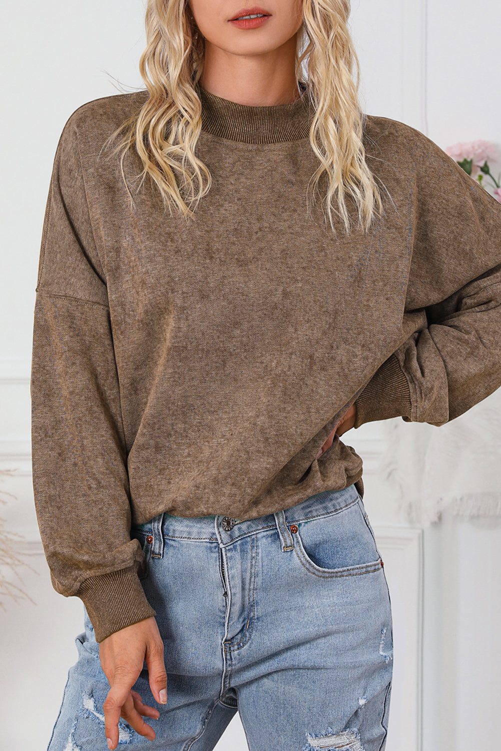 A person is seen wearing a brown Drop Shoulder Crew Neck Pullover Sweatshirt with rolled-up sleeves, paired with light blue jeans and standing with one hand in their pocket. Multiple bracelets are visible on their wrist while the background appears blurred.