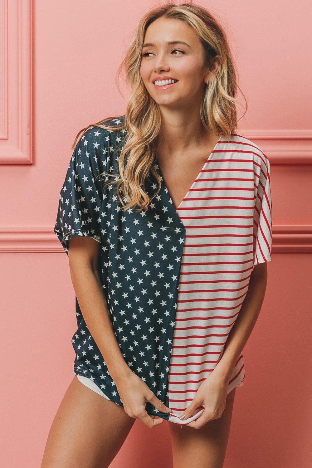 A woman stands against a pink wall, wearing the BiBi US Flag Themed Color Block Short Sleeve T-Shirt, which features one half blue with white stars and the other half red and white stripes. She is smiling with long wavy hair, showcasing her American pride casual wear.