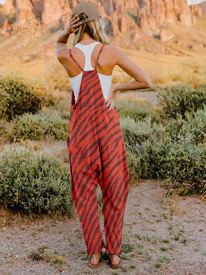 A person wearing a casual white V-neck tank top and a Full Size Printed V-Neck Sleeveless Jumpsuit with a brown animal print zebra-stripe pattern stands outdoors with one hand on their hat and the other in their pocket, set against a rocky landscape backdrop.