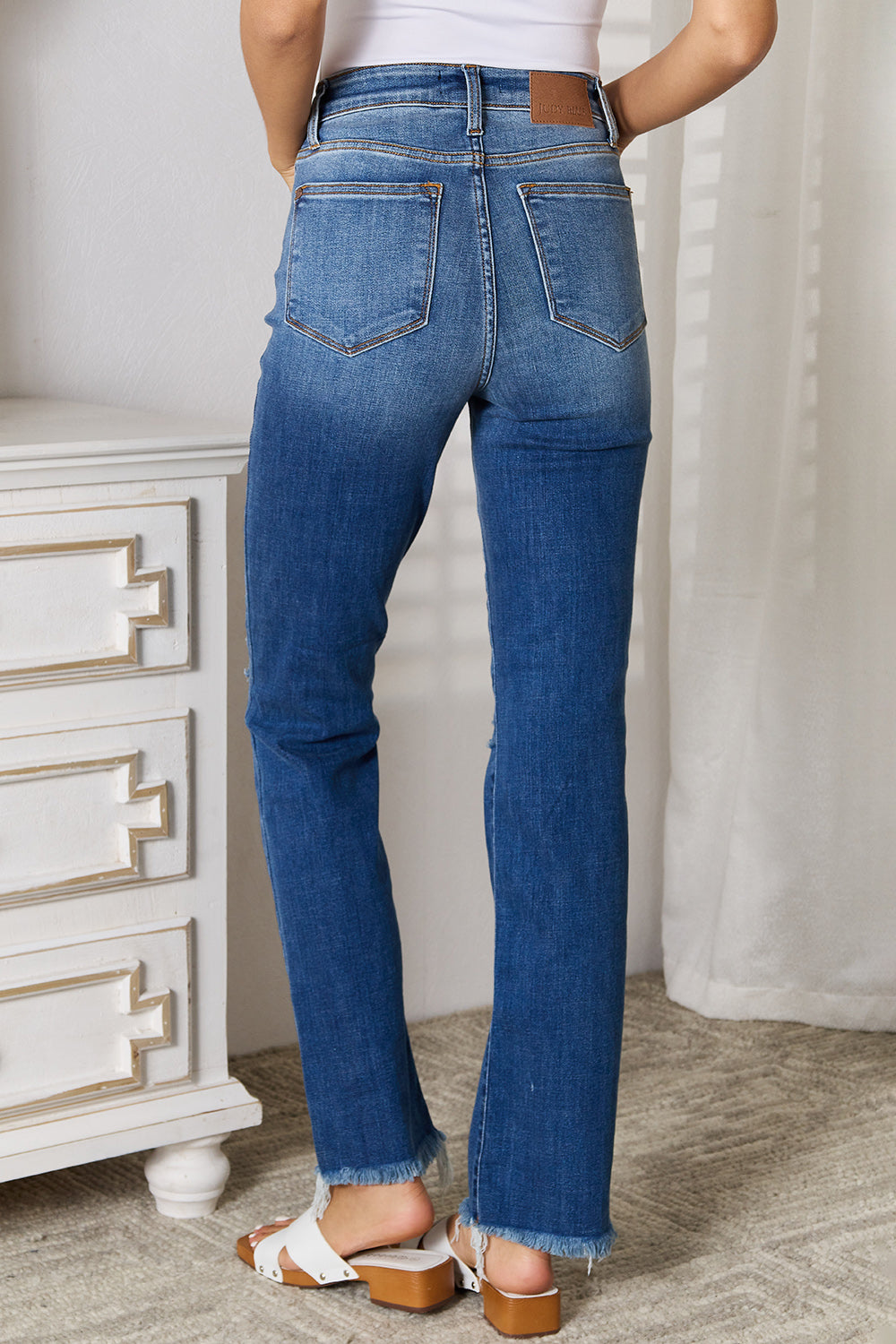 A person wearing Judy Blue Full Size Distressed Raw Hem Jeans, which are slightly stretchy and blue, along with a white top stands indoors near a white dresser.