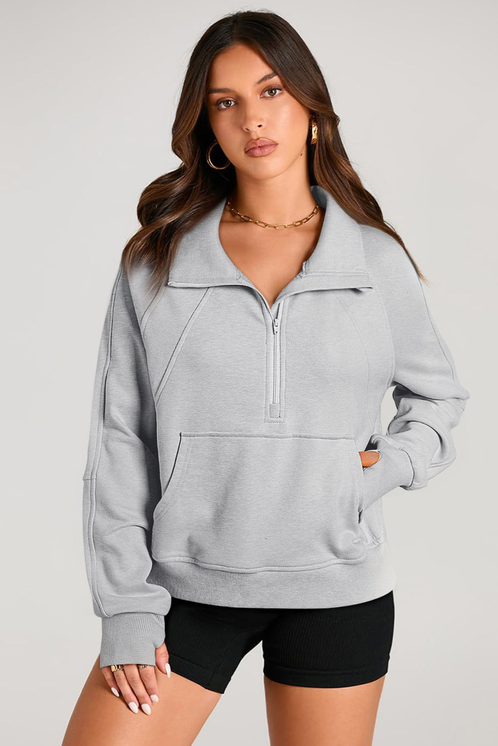 A woman with long, dark hair stands sideways, wearing a Light Grey Quarter Zip Stand Neck Kangaroo Pocket Sweatshirt that is perfect for layering, along with black shorts. She looks at the camera against a plain background, making this outfit ideal for casual occasions.