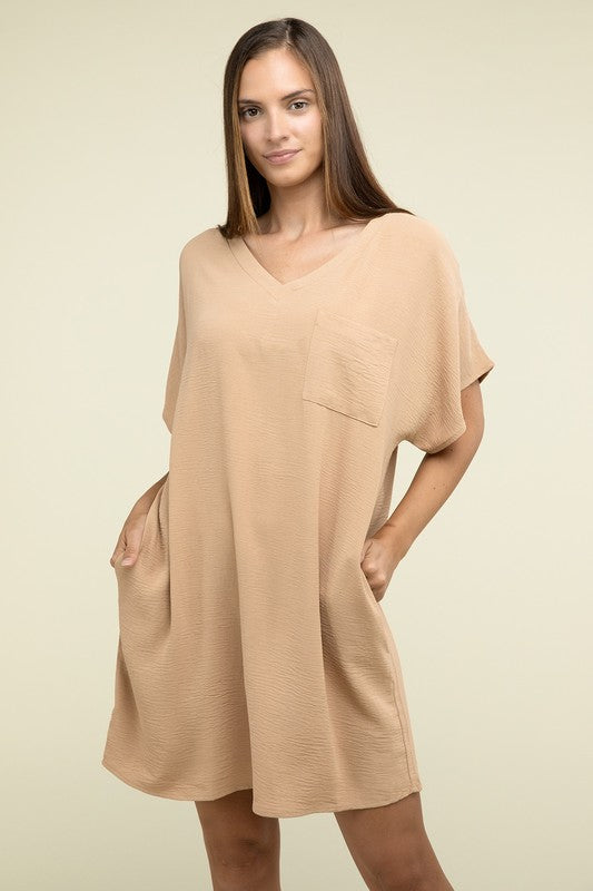 A woman wearing a loose light brown Woven Airflow V Neck T-Shirt Dress with Pockets stands against a plain background. Made from breathable fabric, this summer dress includes pockets where her hands rest as she smiles slightly.