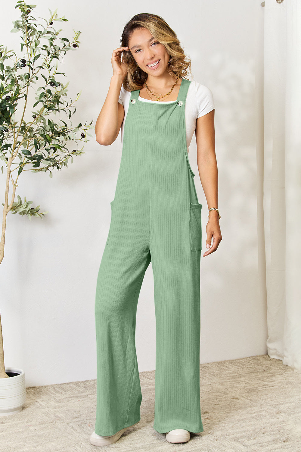 A smiling person wearing the "Double Take Full Size Wide Strap Overall with Pockets," known for its basic style and size-inclusive fit, stands indoors next to a potted plant.