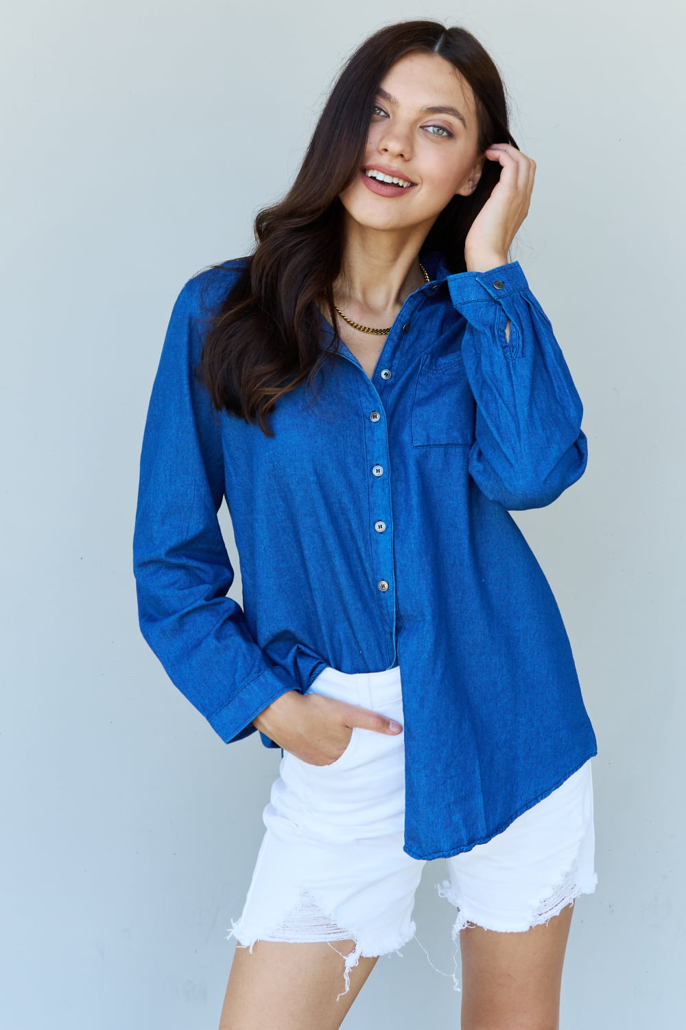 A person standing against a plain background, wearing the Doublju Blue Jean Baby Denim Button Down Shirt Top in Dark Blue and white shorts, smiling and holding a hand to their hair.