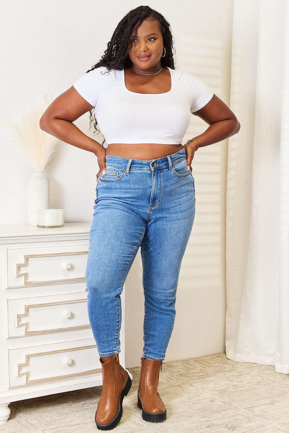 A person wearing Judy Blue Full Size High Waist Skinny Jeans in medium ash wash with a flattering fit, a white top, and brown boots stands near a white dresser, casually slipping one hand into their pocket.