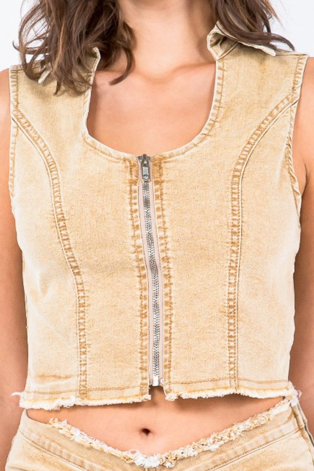 A woman with brunette hair wears an American Bazi Zip Up Washed Crop Denim Vest, a slim-fitting sleeveless cropped top with a front zipper closure, paired with matching high-waisted denim pants.