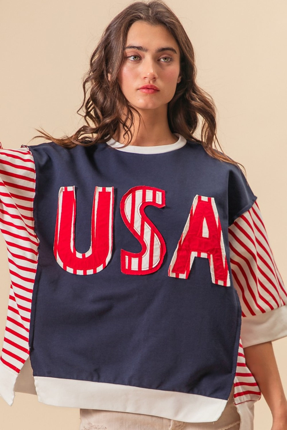 A person dressed in a “BiBi USA Letter Patchwork Contrast Short Sleeve T-Shirt” featuring blue, red, and white colors with striped sleeves, paired with beige shorts, poses against a beige background, proudly displaying their American spirit.