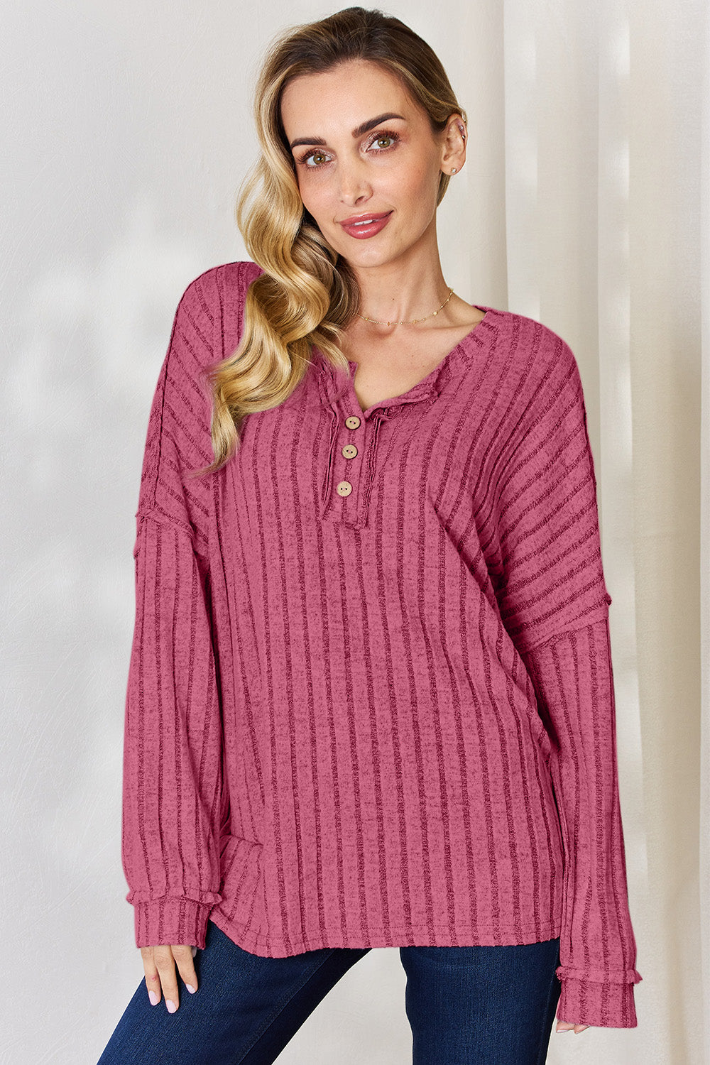 A woman with long, wavy blonde hair wears a slightly stretchy, textured Basic Bae Full Size Ribbed Half Button Long Sleeve T-Shirt in pink, standing against a neutral backdrop.