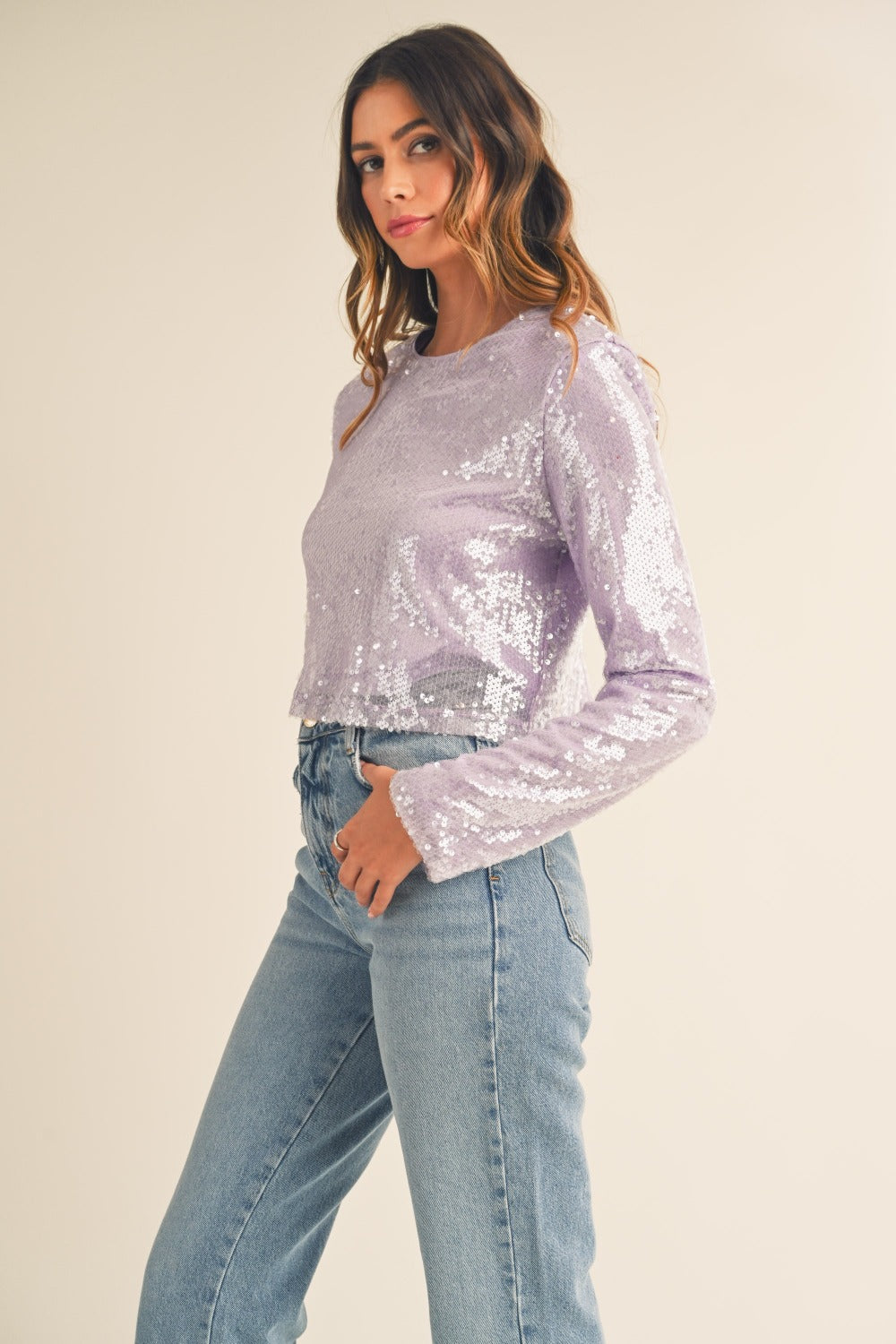 A woman with long, wavy hair wears a trendy MABLE Shoulder Padded Sequin Crop Top in light purple along with light blue jeans, standing against a plain off-white background.